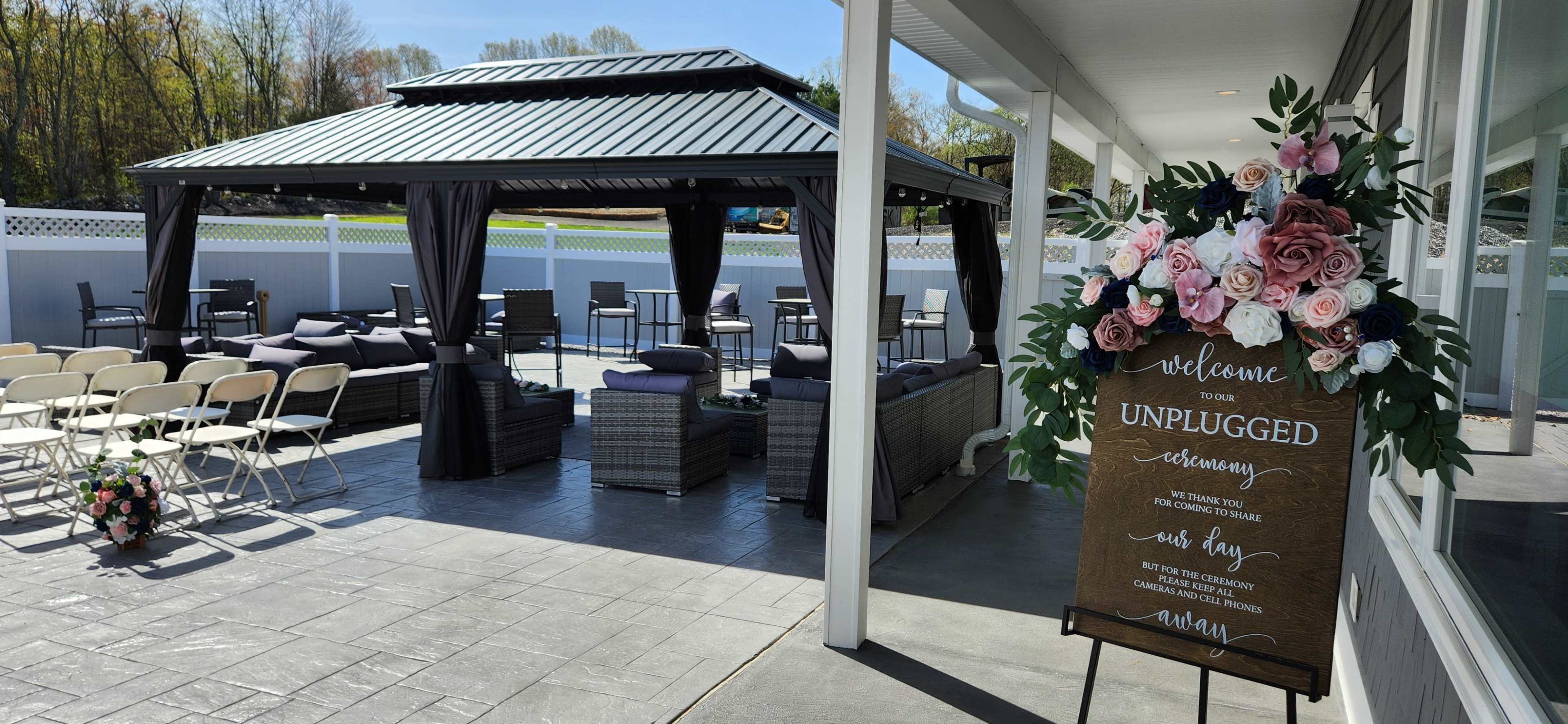 The scene shows an outdoor event space with seating arranged in front of a decorated gazebo, featuring a welcome sign and floral accents.