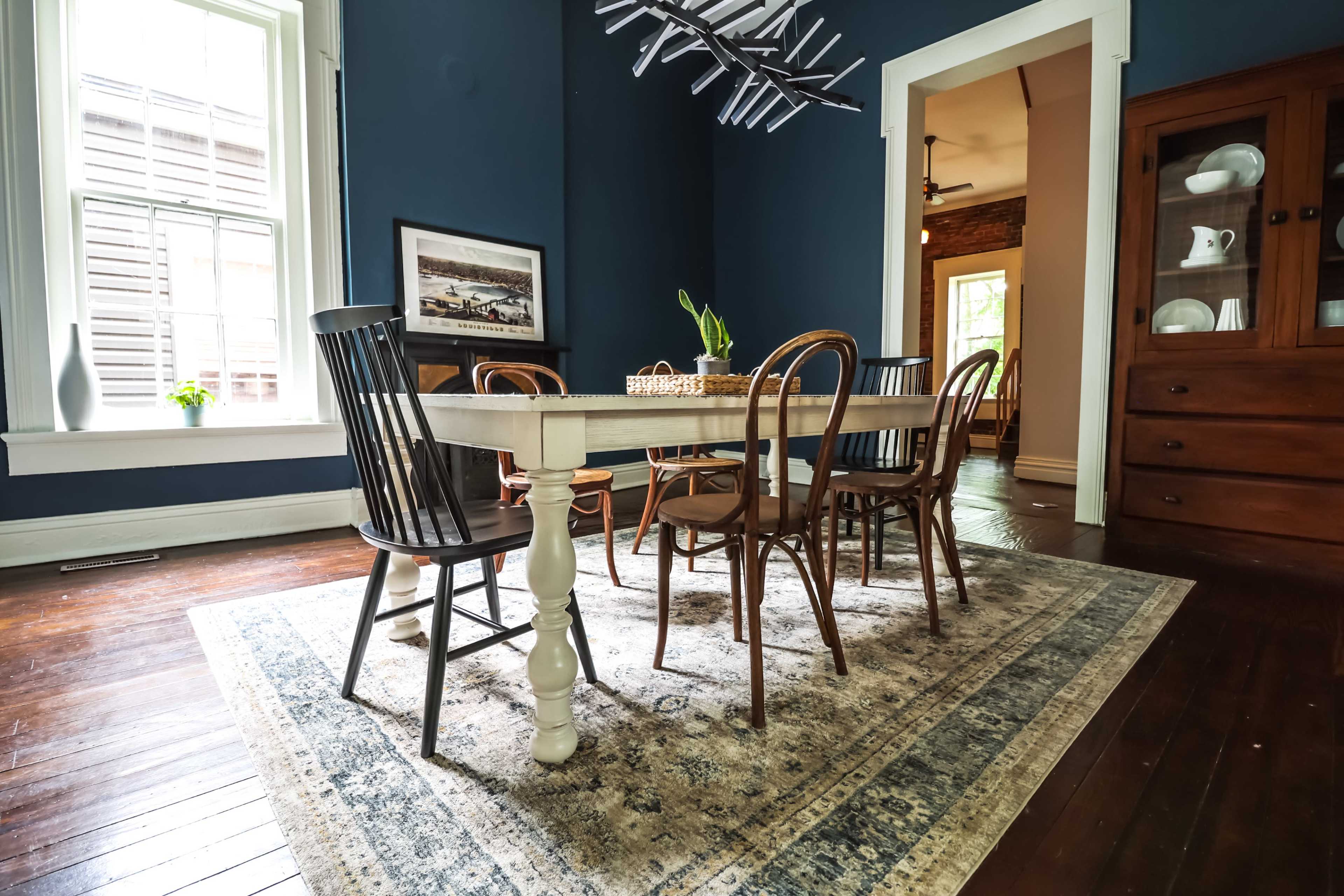 A dining table with five chairs is set in a room featuring dark blue walls and hardwood flooring, with a decorative rug underneath.