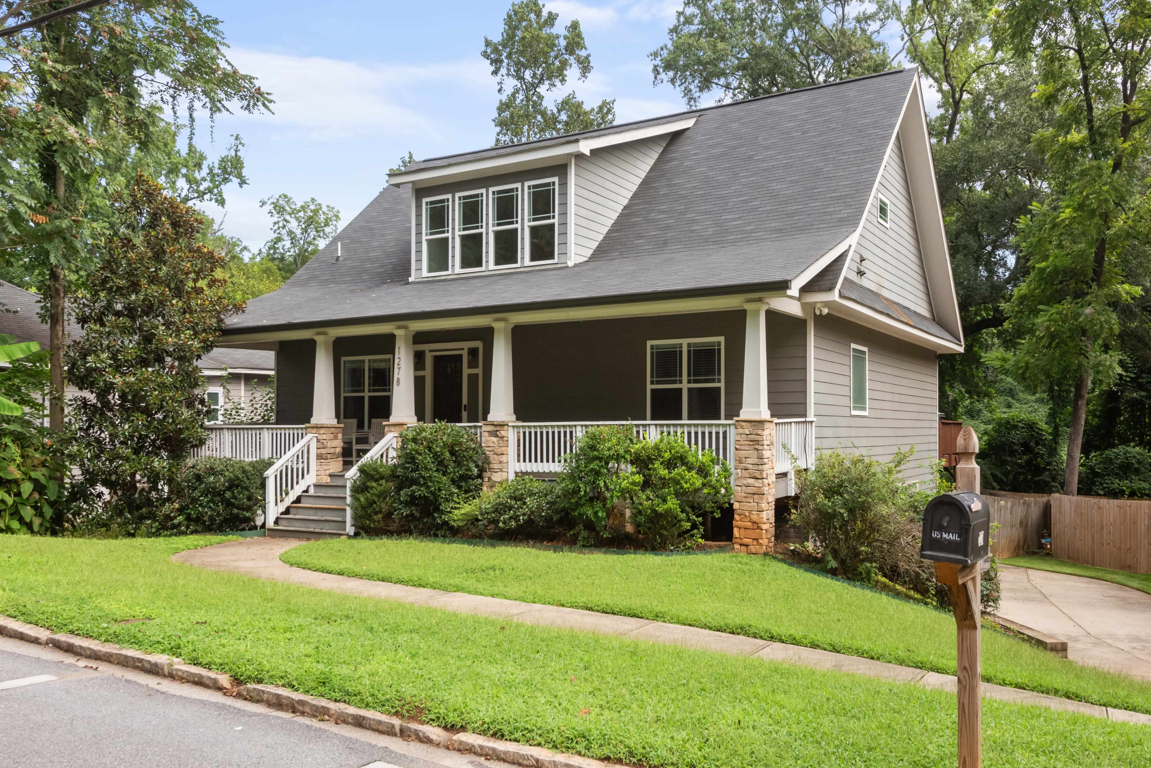 A two-story house with a grey exterior and white trim features a front porch and is surrounded by green landscaping.