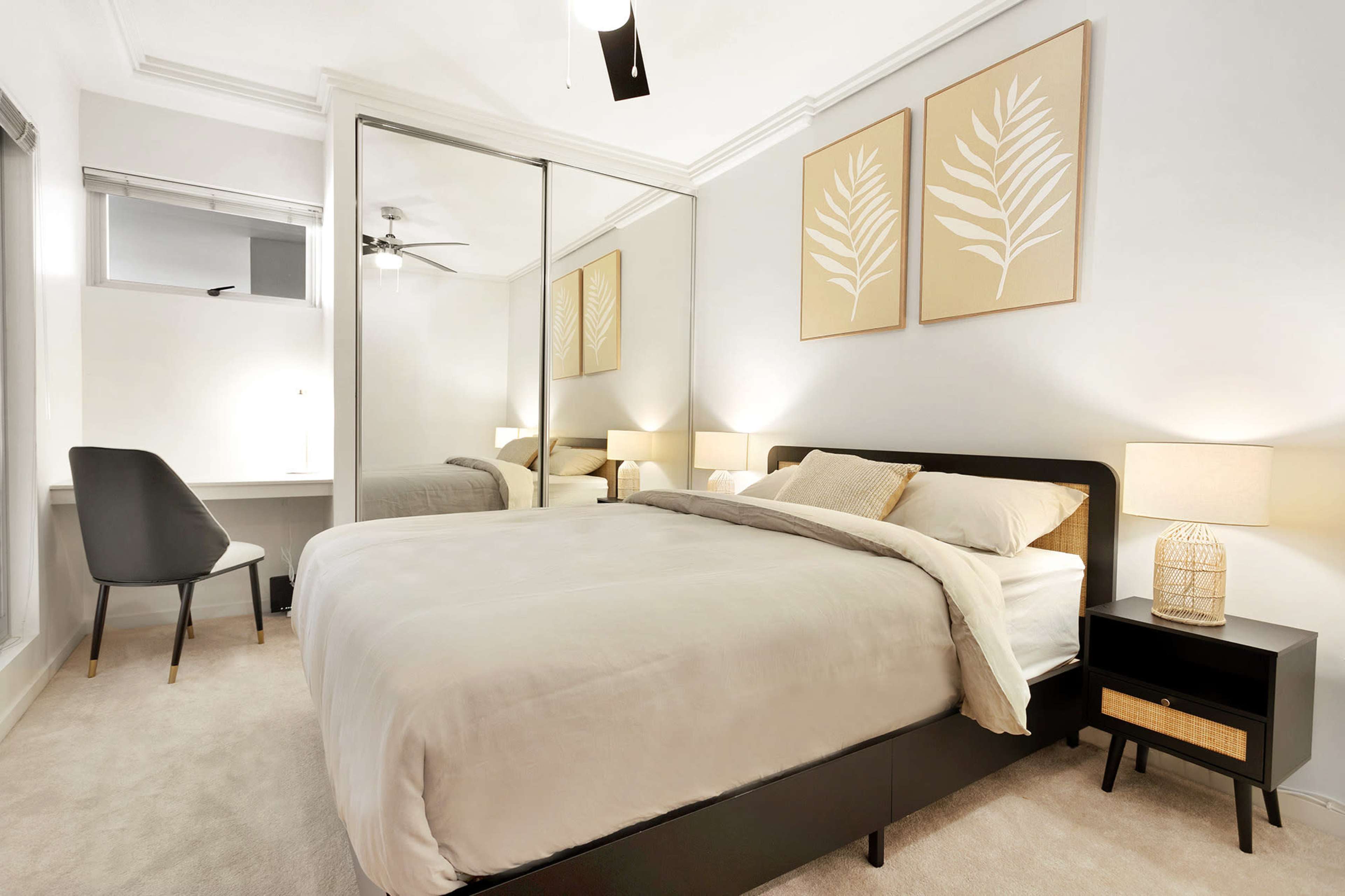 The image shows a modern bedroom with a bed, nightstand, and desk, featuring a large mirror and two framed leaf prints on the wall.