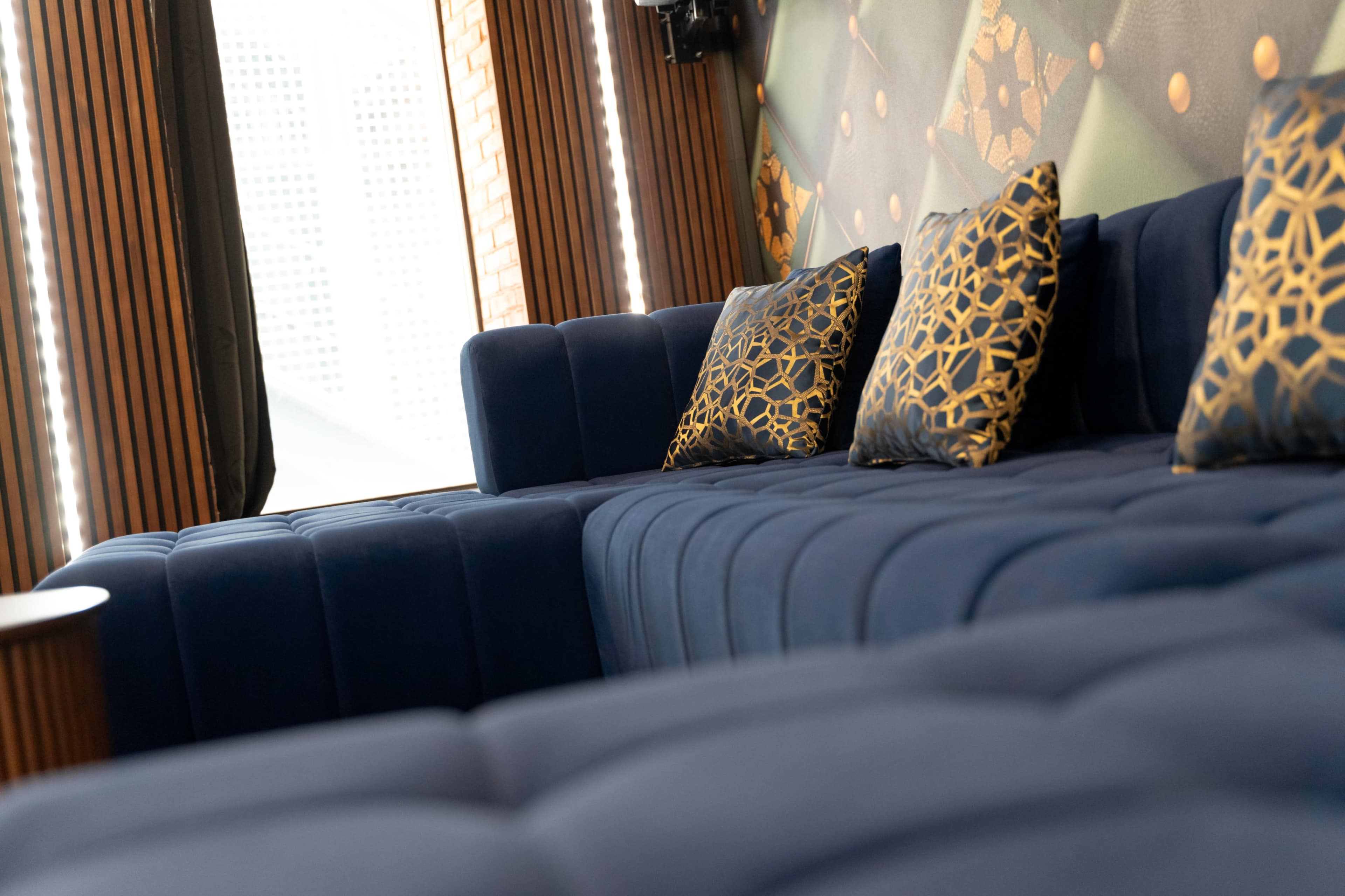 The image shows a modern lounge area featuring a blue velvet sectional sofa adorned with decorative gold-patterned cushions.