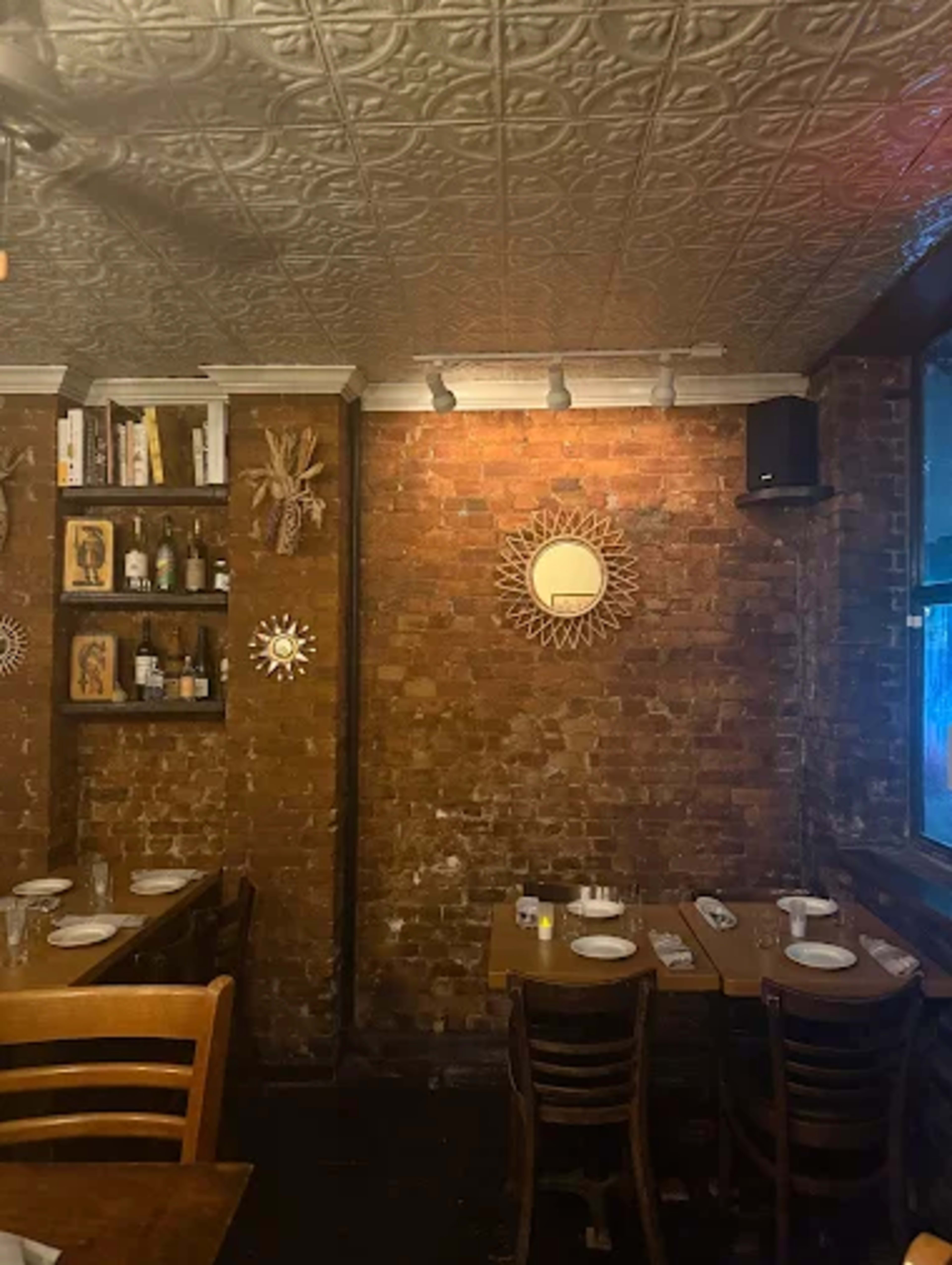 The image shows a cozy restaurant interior with exposed brick walls, wooden tables, and shelving displaying bottles and decorative items.