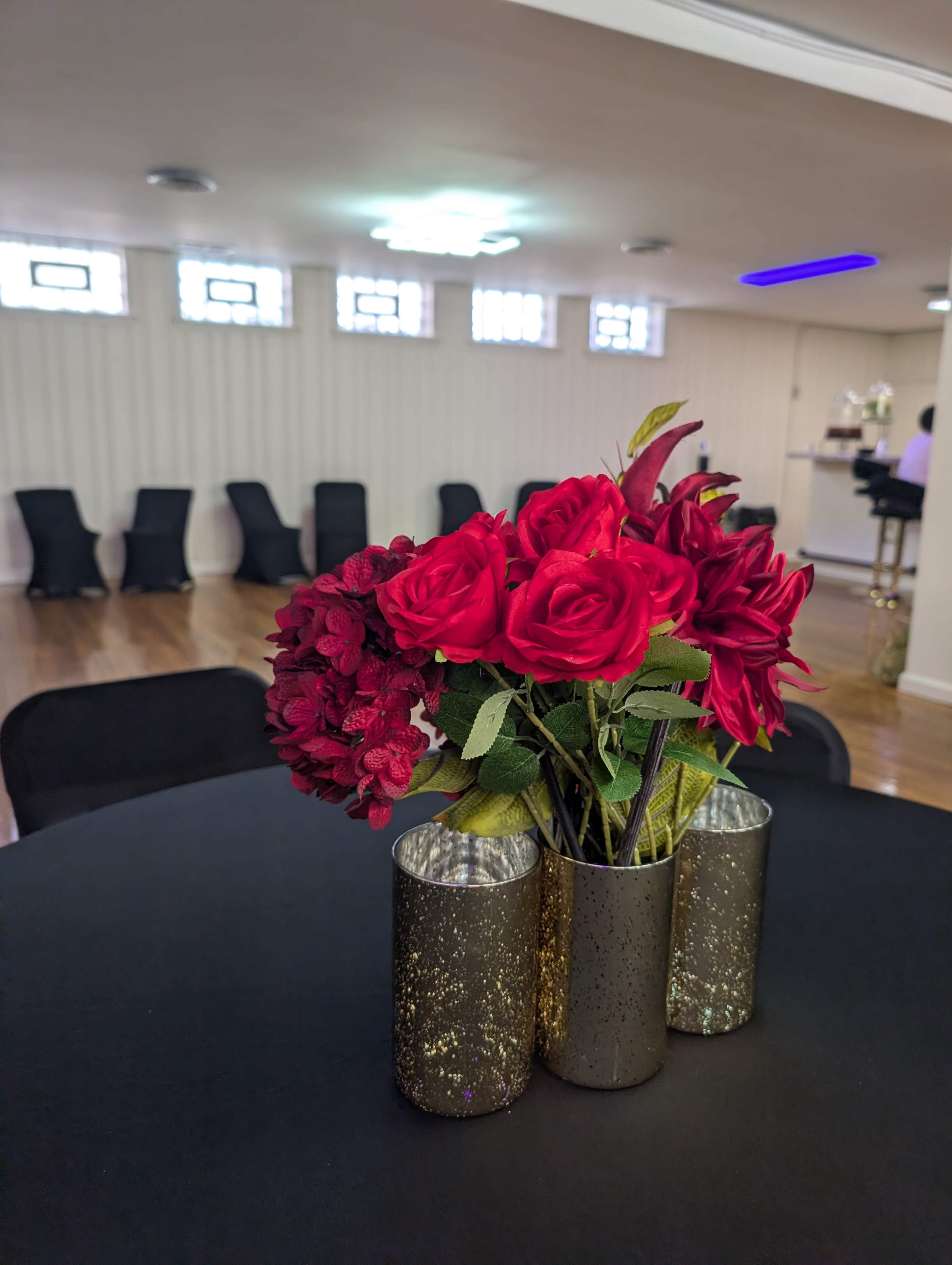 A bouquet of red artificial roses is placed in a glittery vase on a black table, with chairs and a bar area visible in the background.