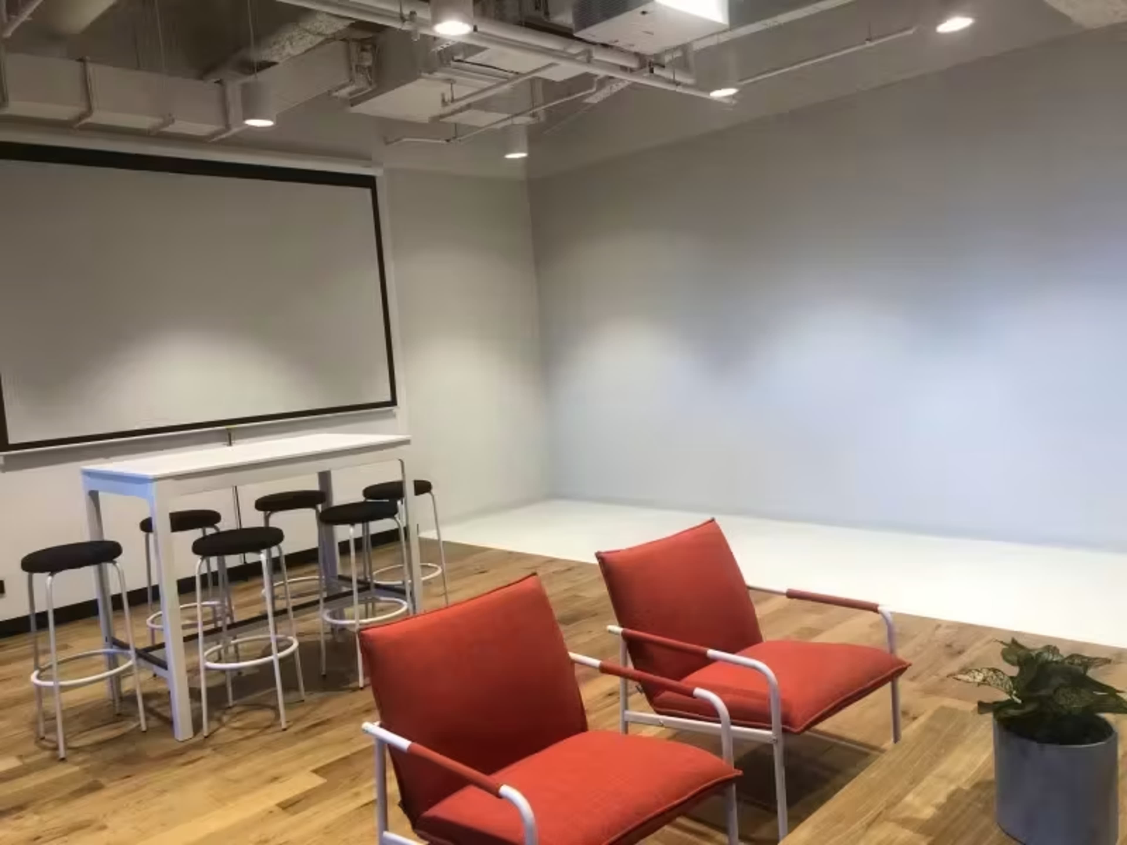 A modern office space featuring two red chairs, a high table with stools, and a large projection screen against a light gray wall.