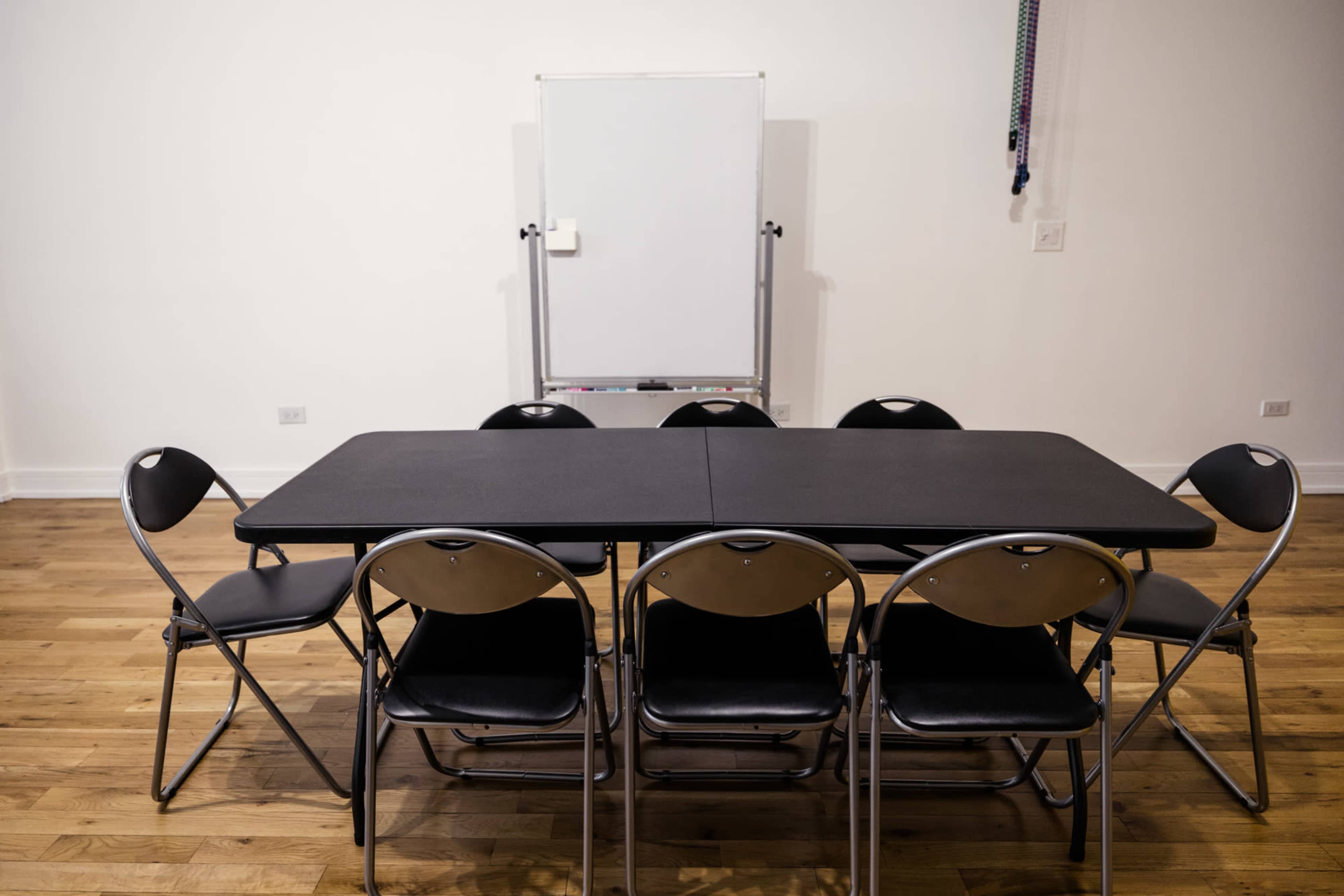 A rectangular table with eight black folding chairs is positioned in front of a blank whiteboard in a simple room with hardwood floors.