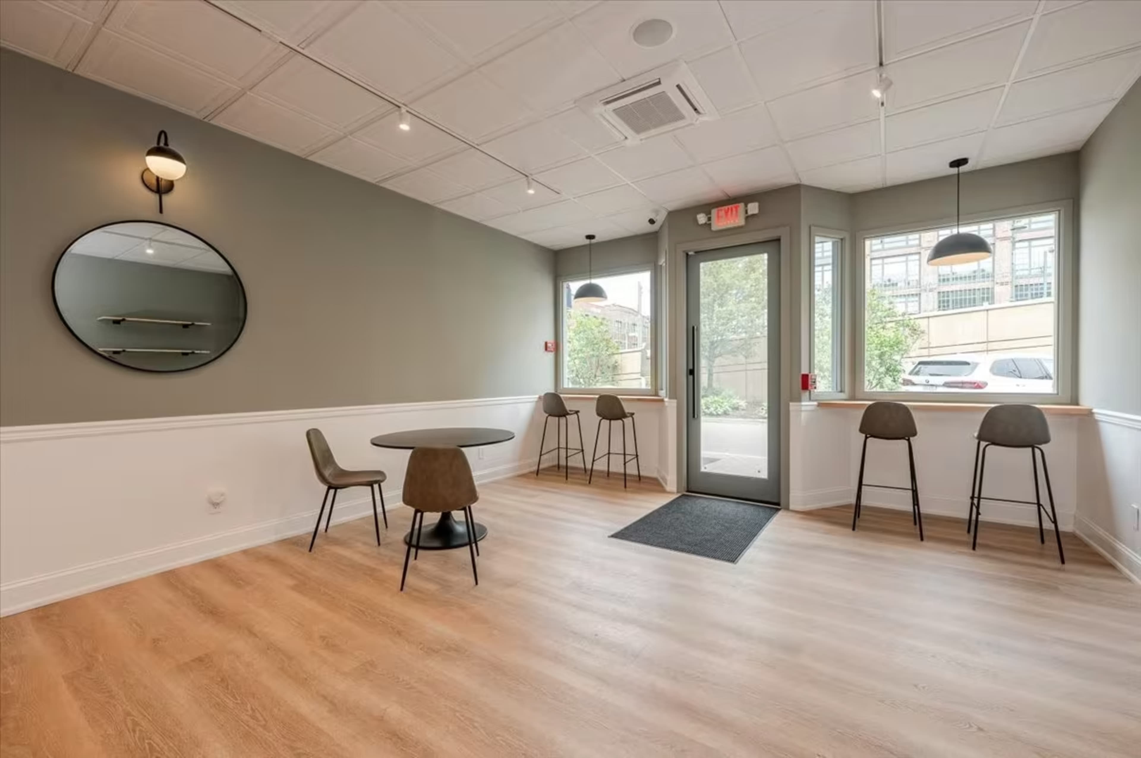 A modern, minimalist interior with a small round table and four chairs, alongside a bar area with three stools, under soft lighting against a muted green wall.