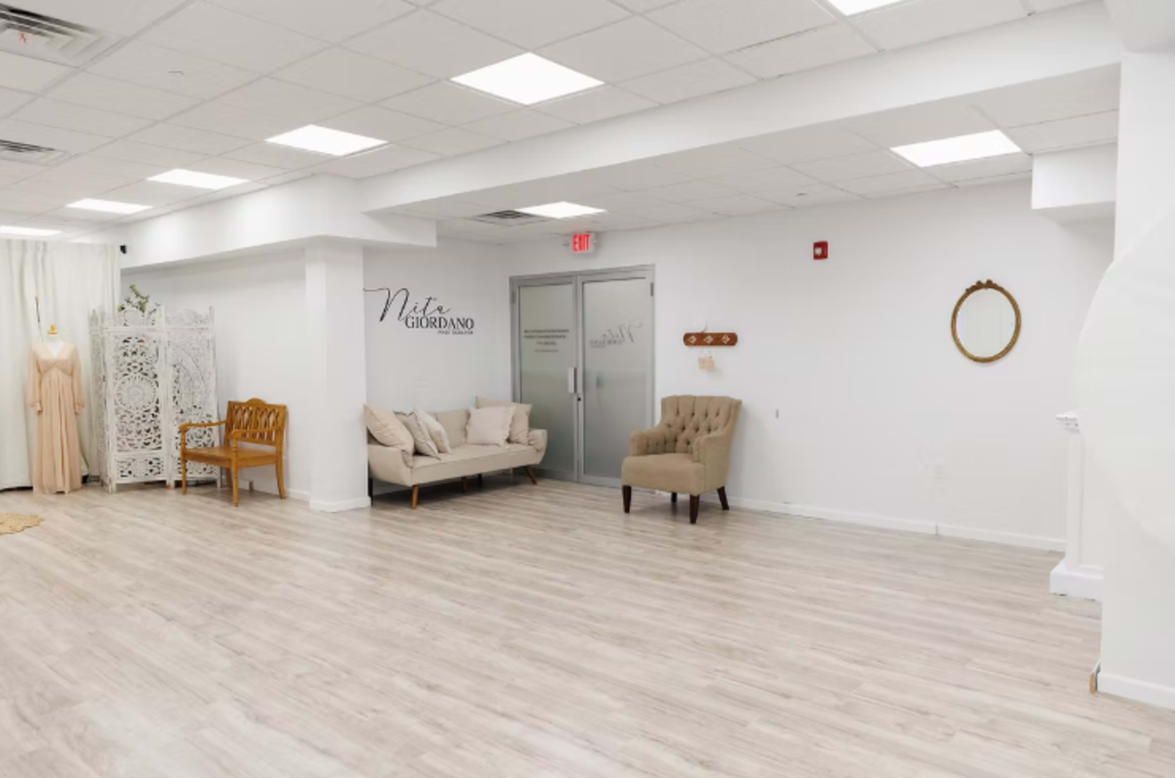 The image shows a spacious, modern room with light-colored wooden flooring, featuring a sofa, an armchair, and decorative wall elements.