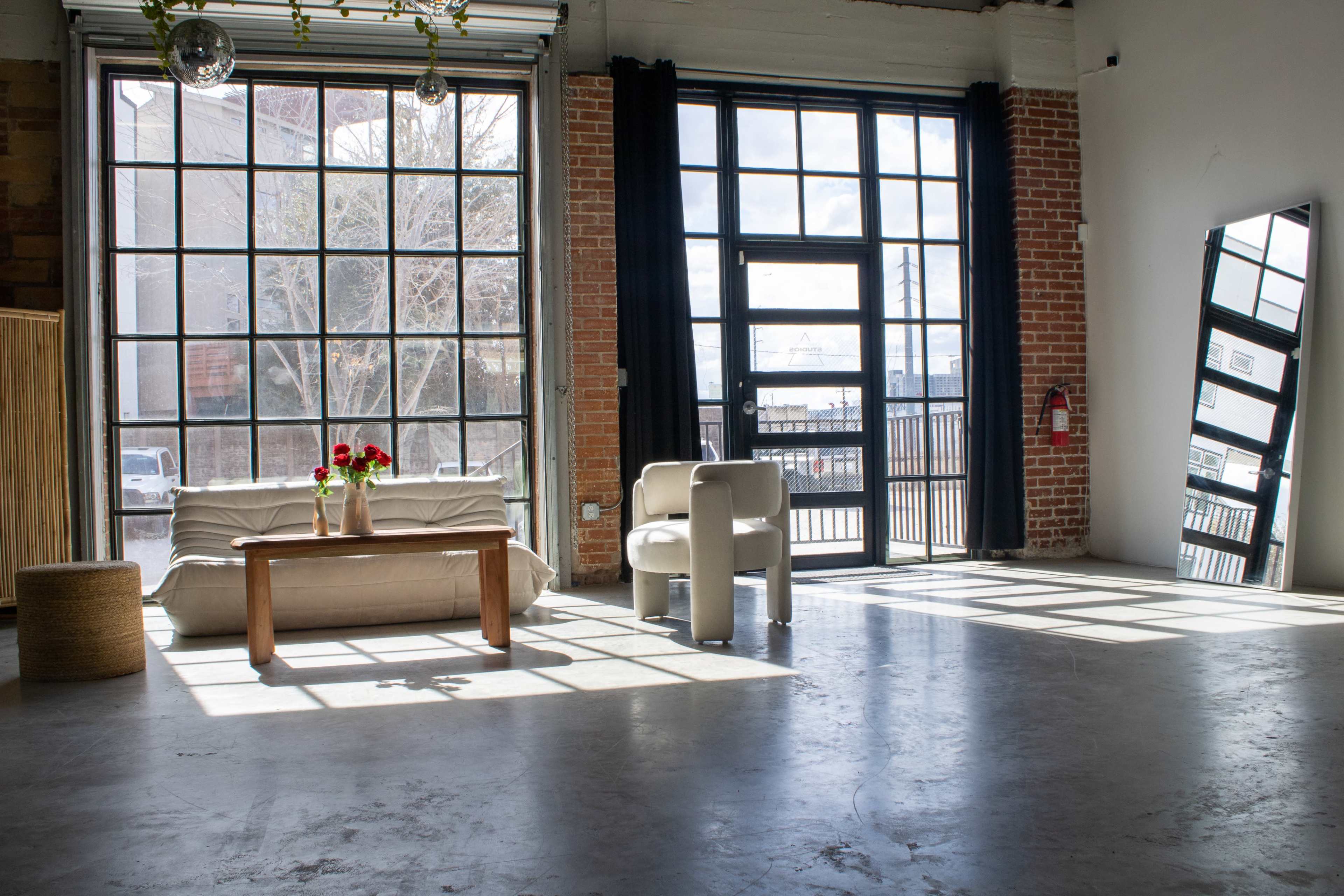 The image shows a spacious, sunlit room with large windows, featuring a white sofa, a wooden coffee table with flowers, and two chairs beside a mirror.