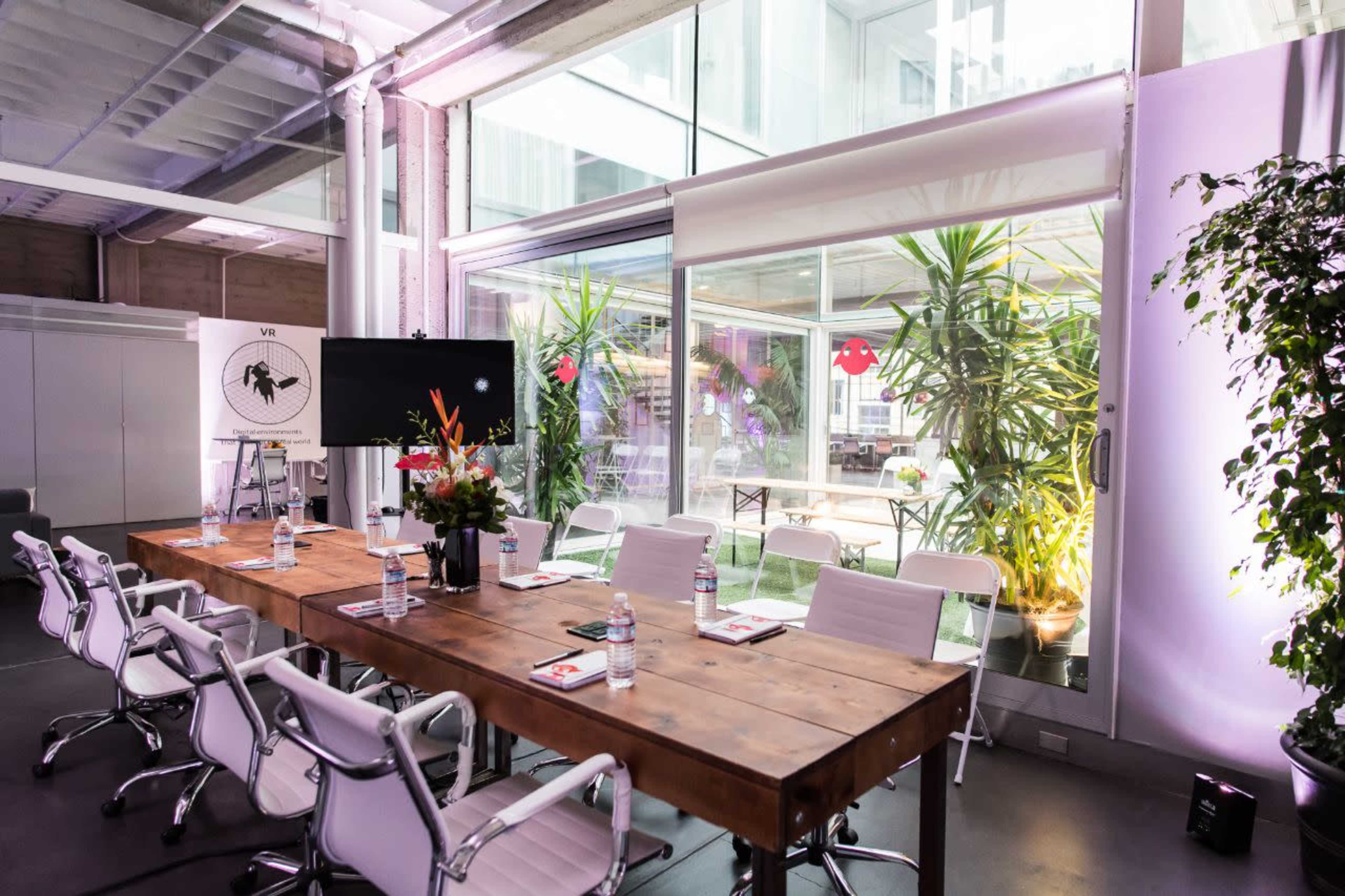 A modern conference room features a long wooden table surrounded by white chairs, with large windows displaying greenery outside.
