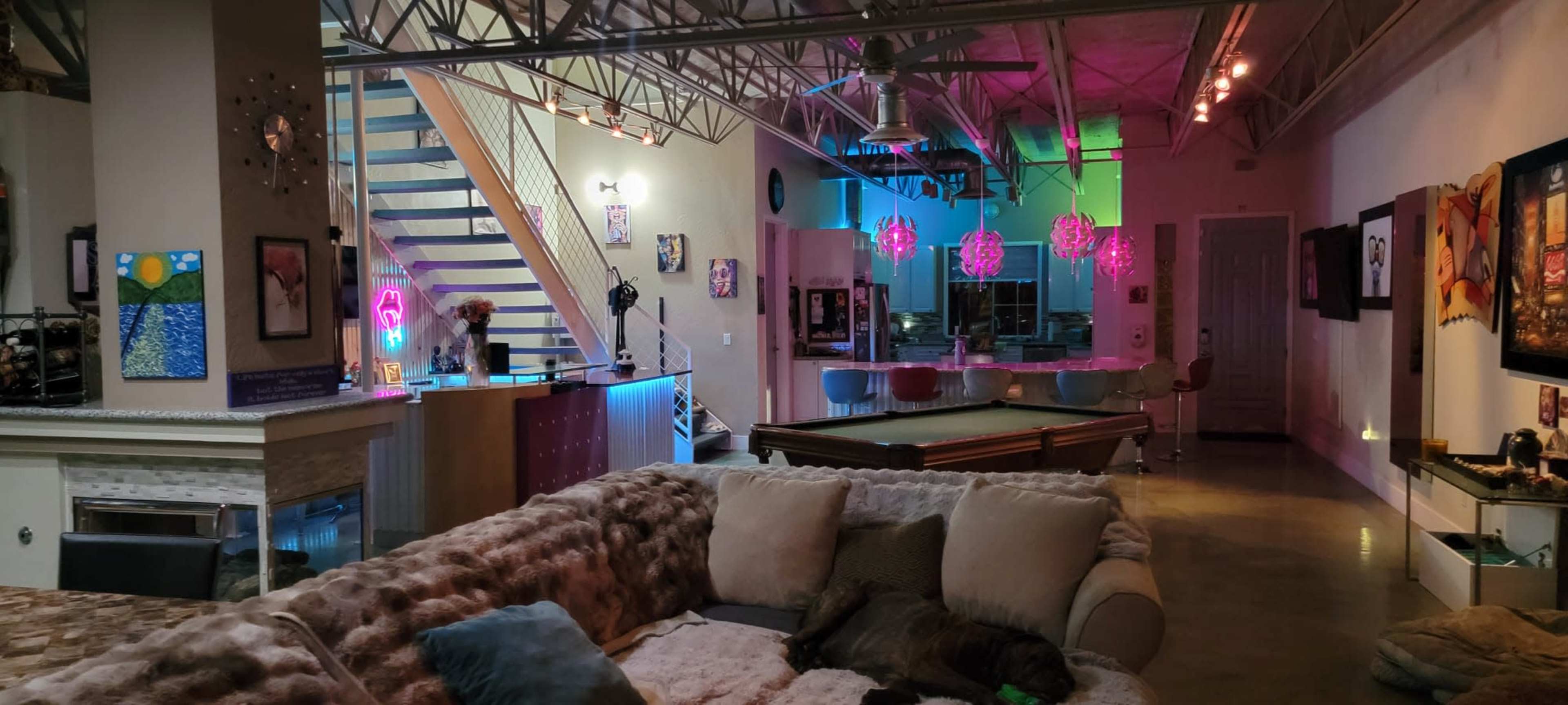 A spacious loft-style living area with a pool table, a large sectional sofa, and colorful lighting from overhead fixtures.