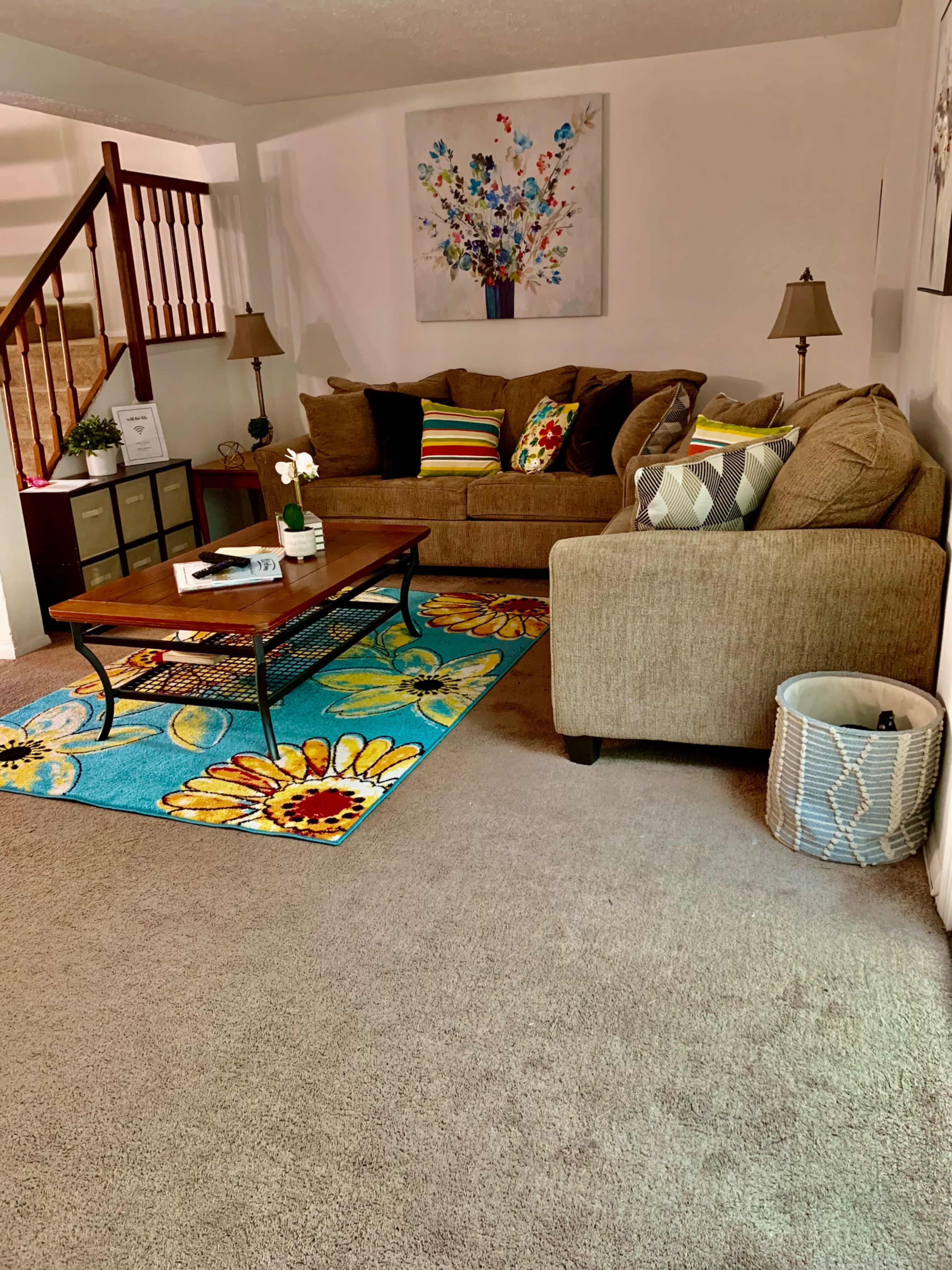 The living room features a brown couch with colorful cushions, a wooden coffee table on a bright floral rug, and a lamp beside the seating area.