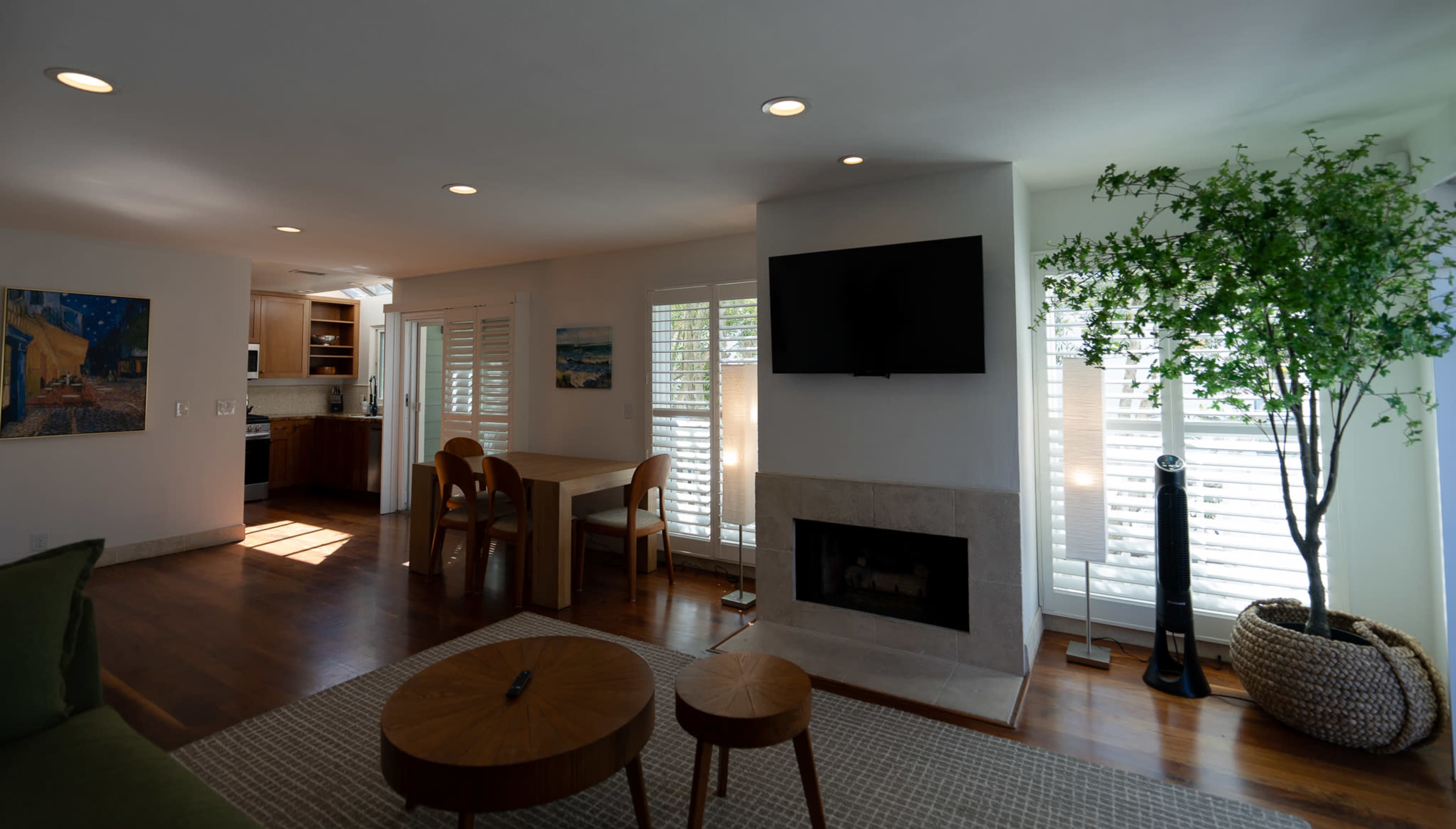 The image shows a modern living room with a fireplace, a television mounted on the wall, a dining table, and natural light coming through window shutters, complemented by a potted plant.