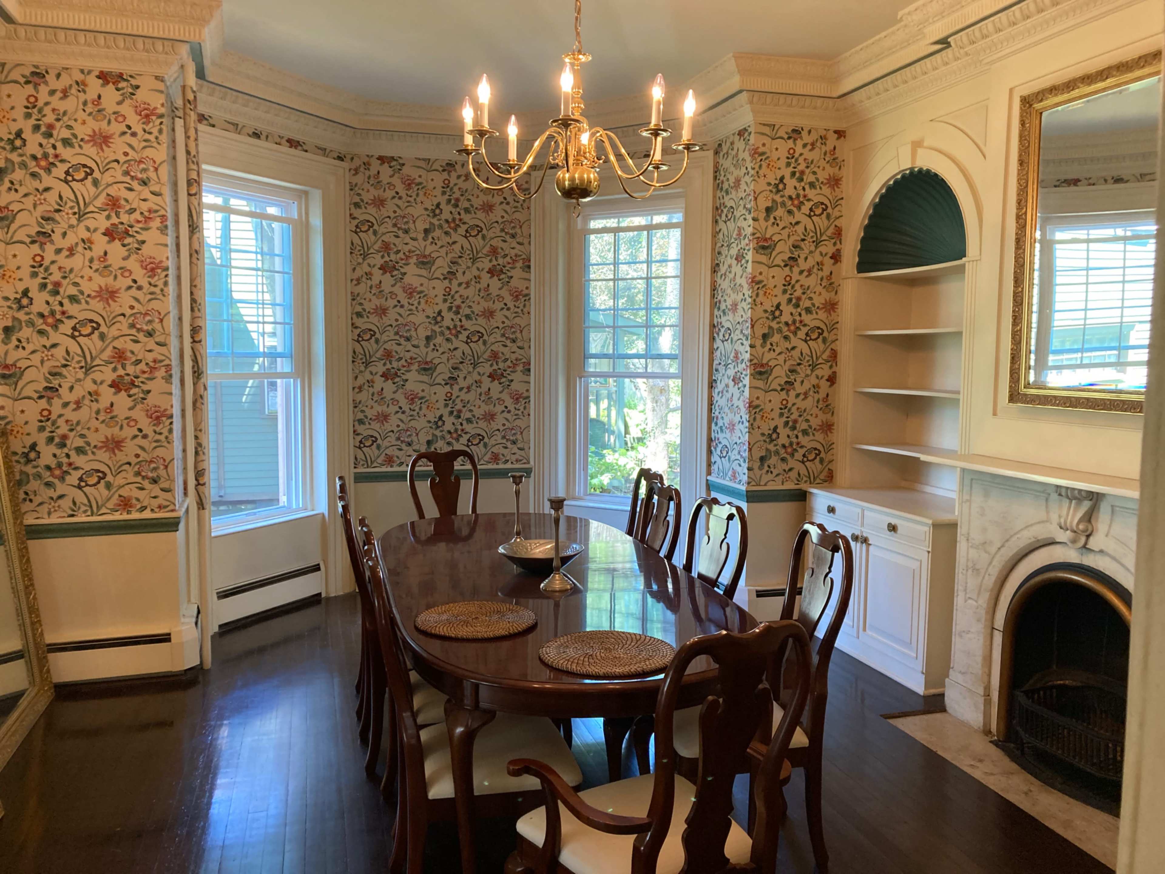 A dining room features a large wooden table surrounded by chairs, with floral wallpaper and a chandelier hanging above.