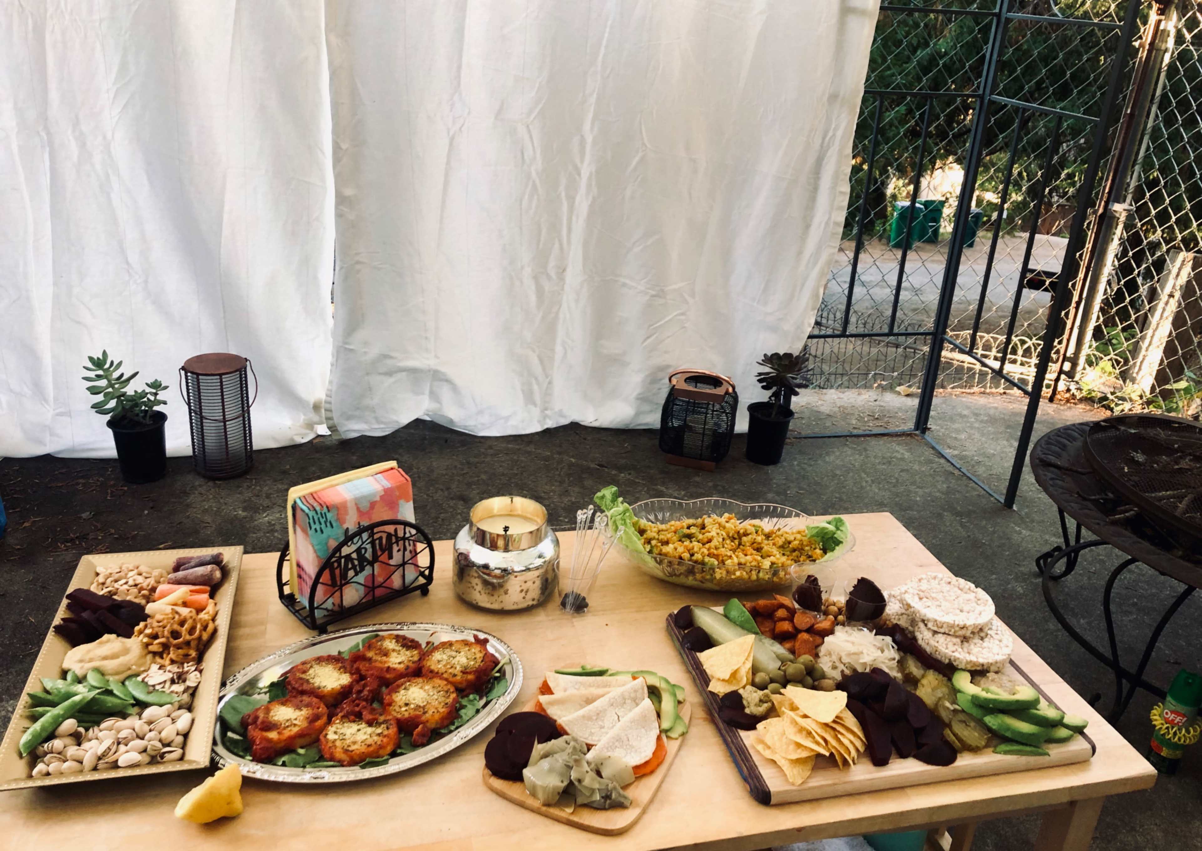 A table is set with a variety of food, including appetizers, a salad, and snacks, against a backdrop of white fabric and greenery.