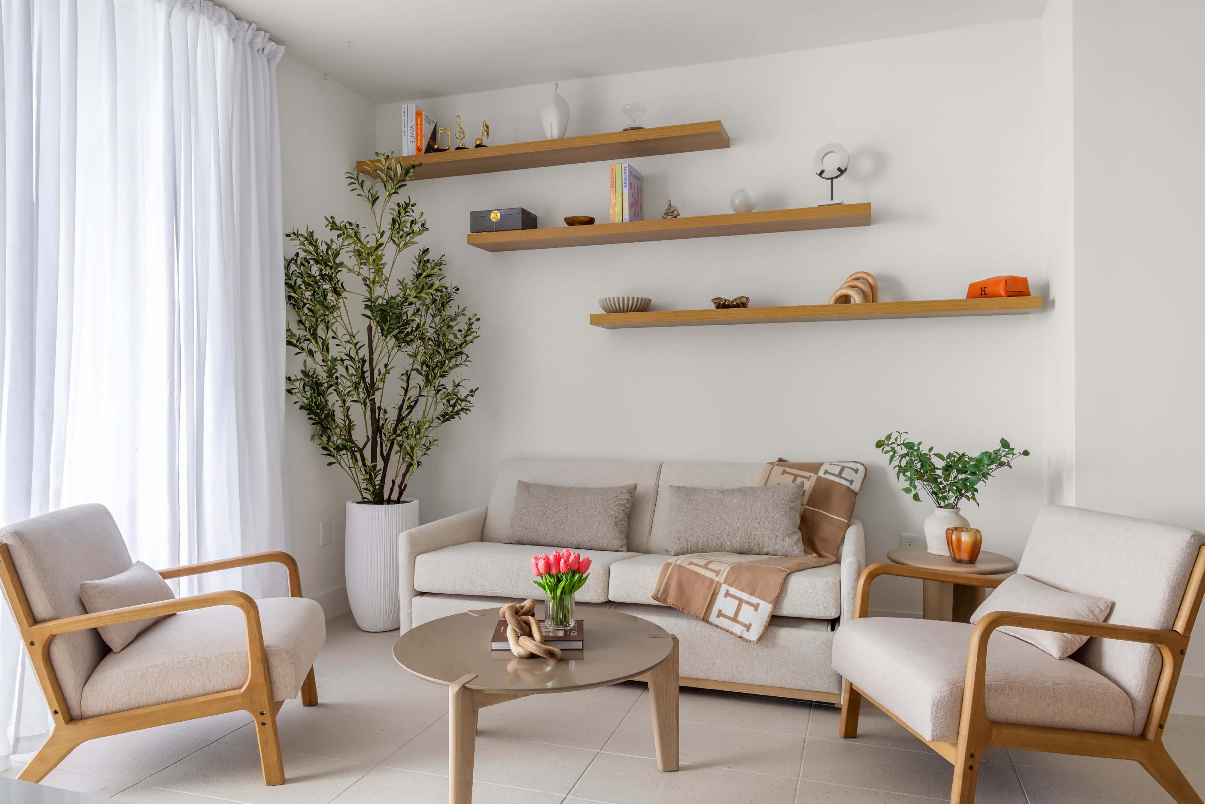 A bright living room features a beige sofa with wooden armchairs, a round table, and floating shelves adorned with decorative items and plants.