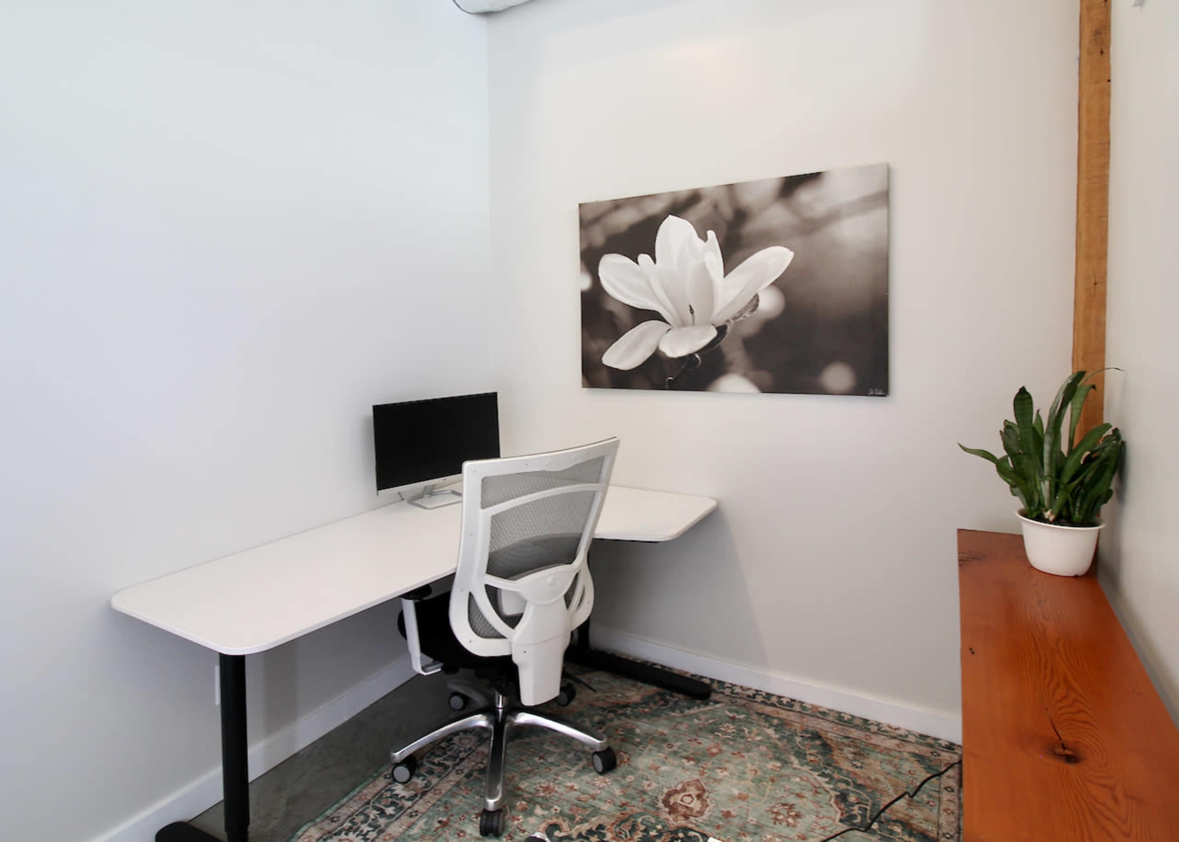 The image shows a minimalistic office space with a white desk, an ergonomic chair, a computer monitor, and a potted plant, set against a wall featuring a black-and-white flower photograph.