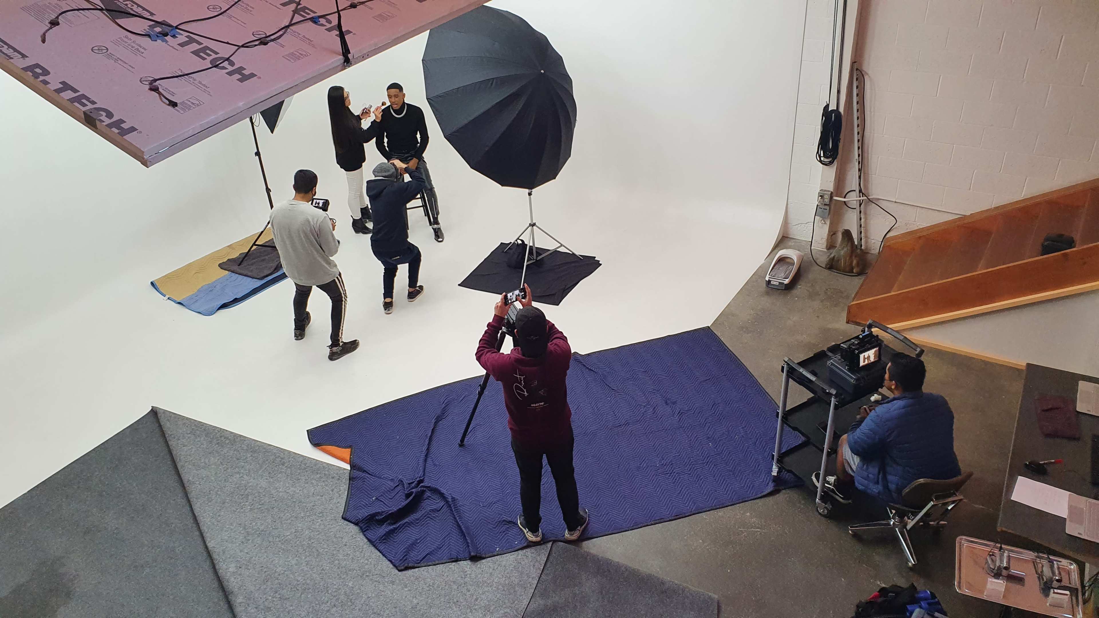 A photography studio is set up with a model sitting on a stool under a large umbrella light, while a photographer captures images and a team assists nearby.