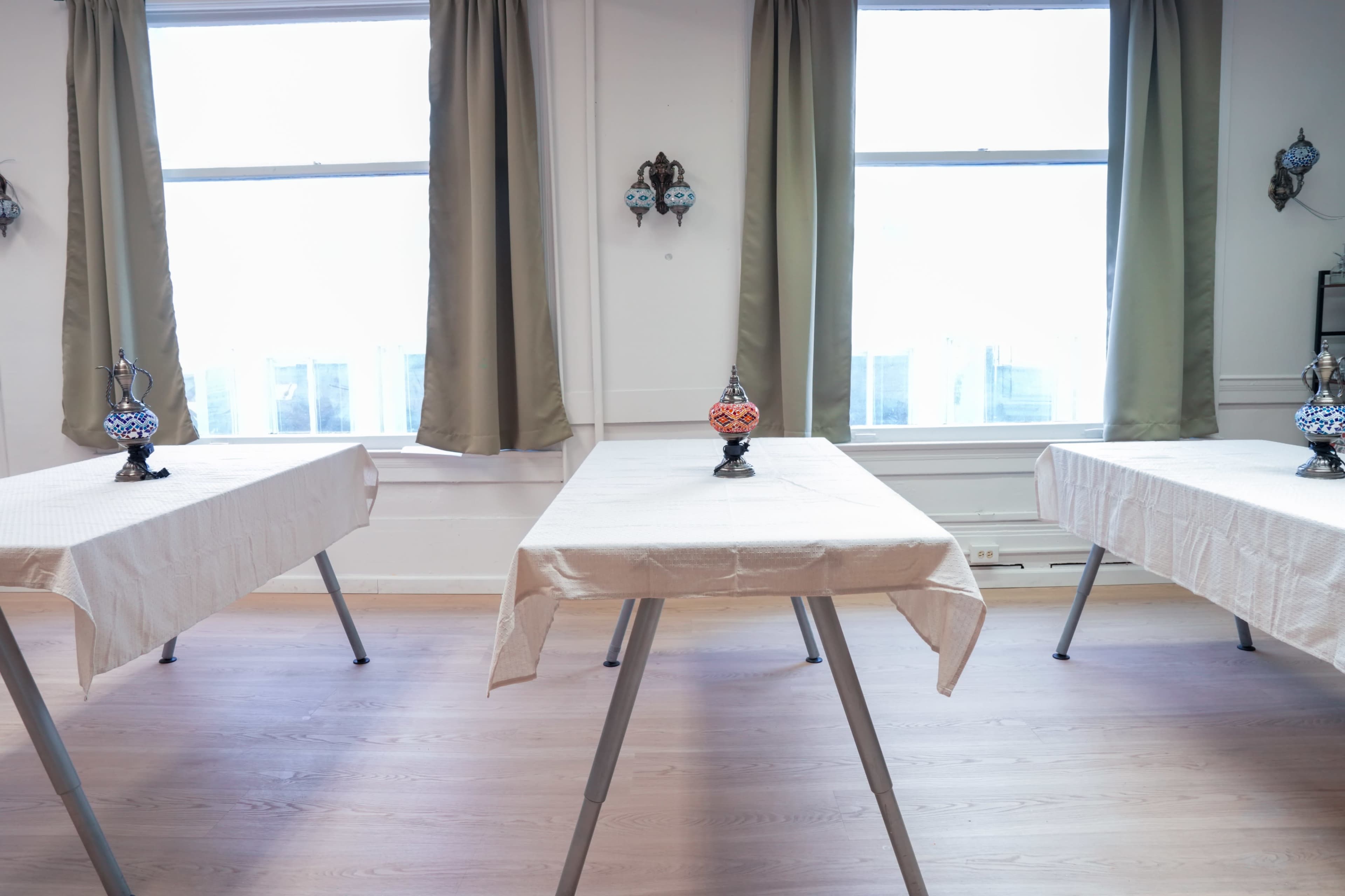 The image shows three tables with tablecloths arranged in a bright room, each decorated with a decorative vase, and large windows letting in natural light.