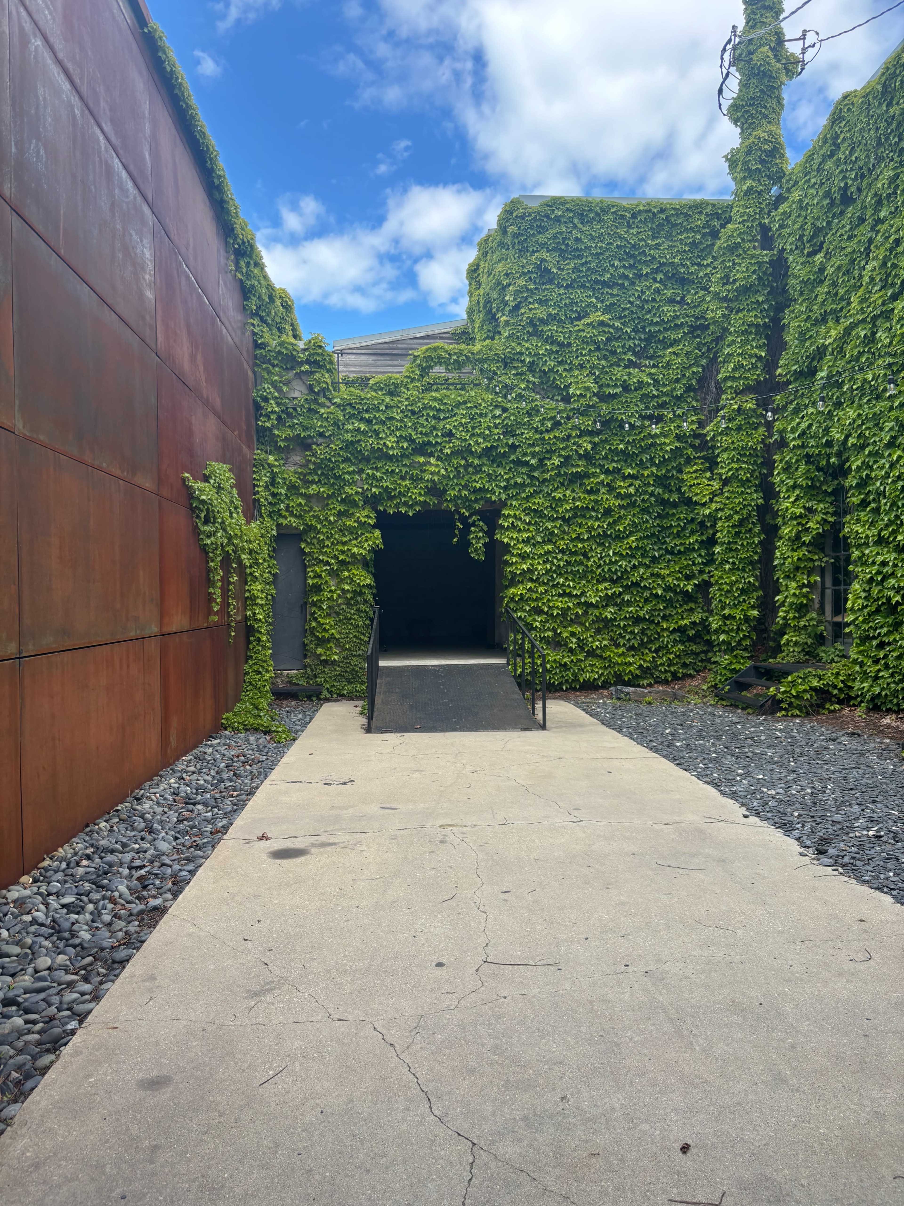 The image shows a pathway leading to an entrance surrounded by walls covered in green ivy and a backdrop of blue sky with clouds.