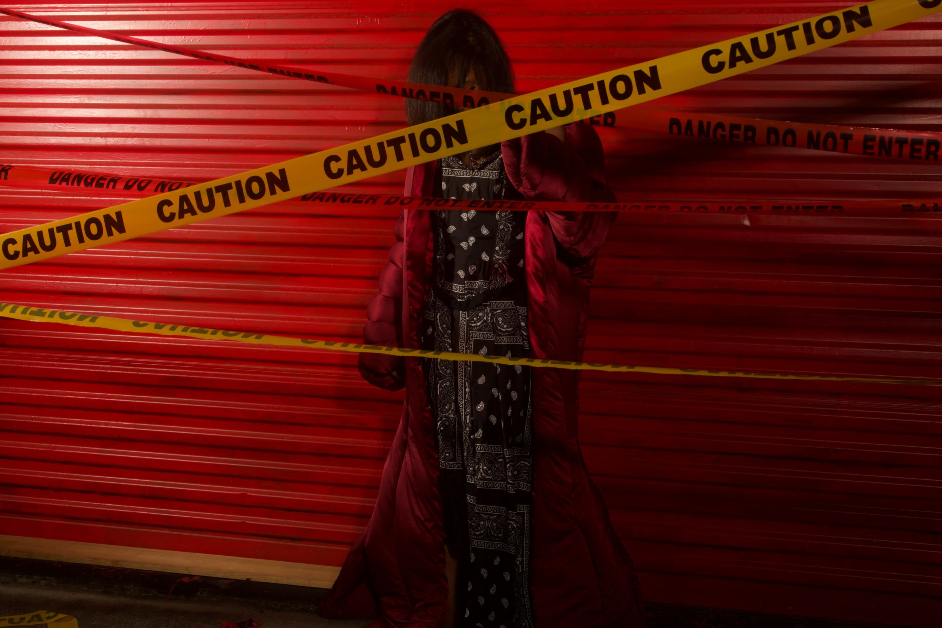 A person wearing a long coat stands behind yellow caution tape in front of a red corrugated metal background.