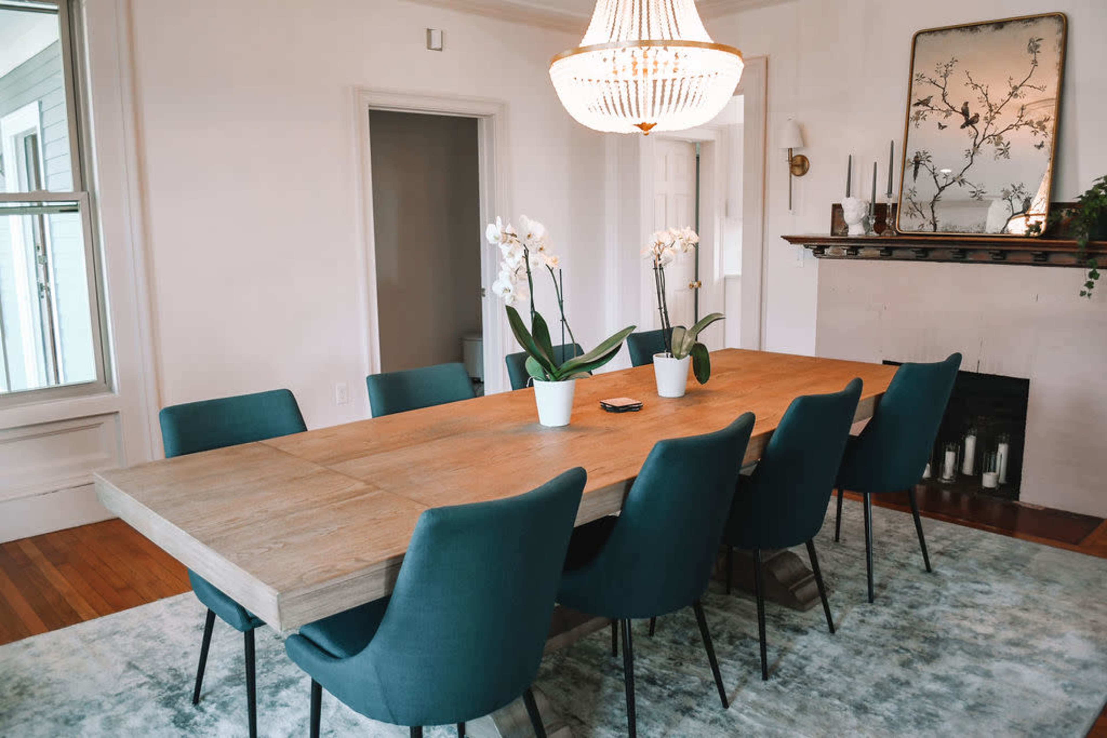The image shows a dining room with a large wooden table surrounded by teal chairs, featuring a chandelier above and potted orchids as centerpieces.