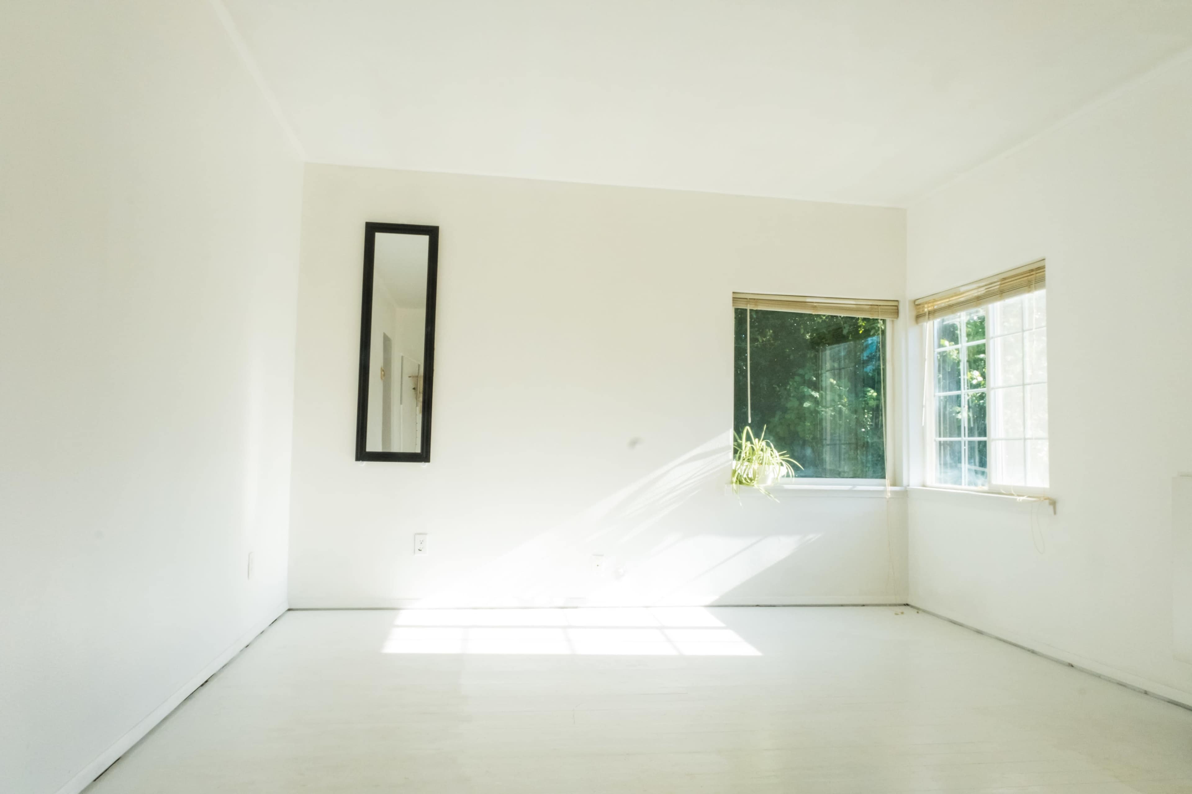 A bright, empty room with white walls, a mirror on one wall, and a window letting in sunlight.