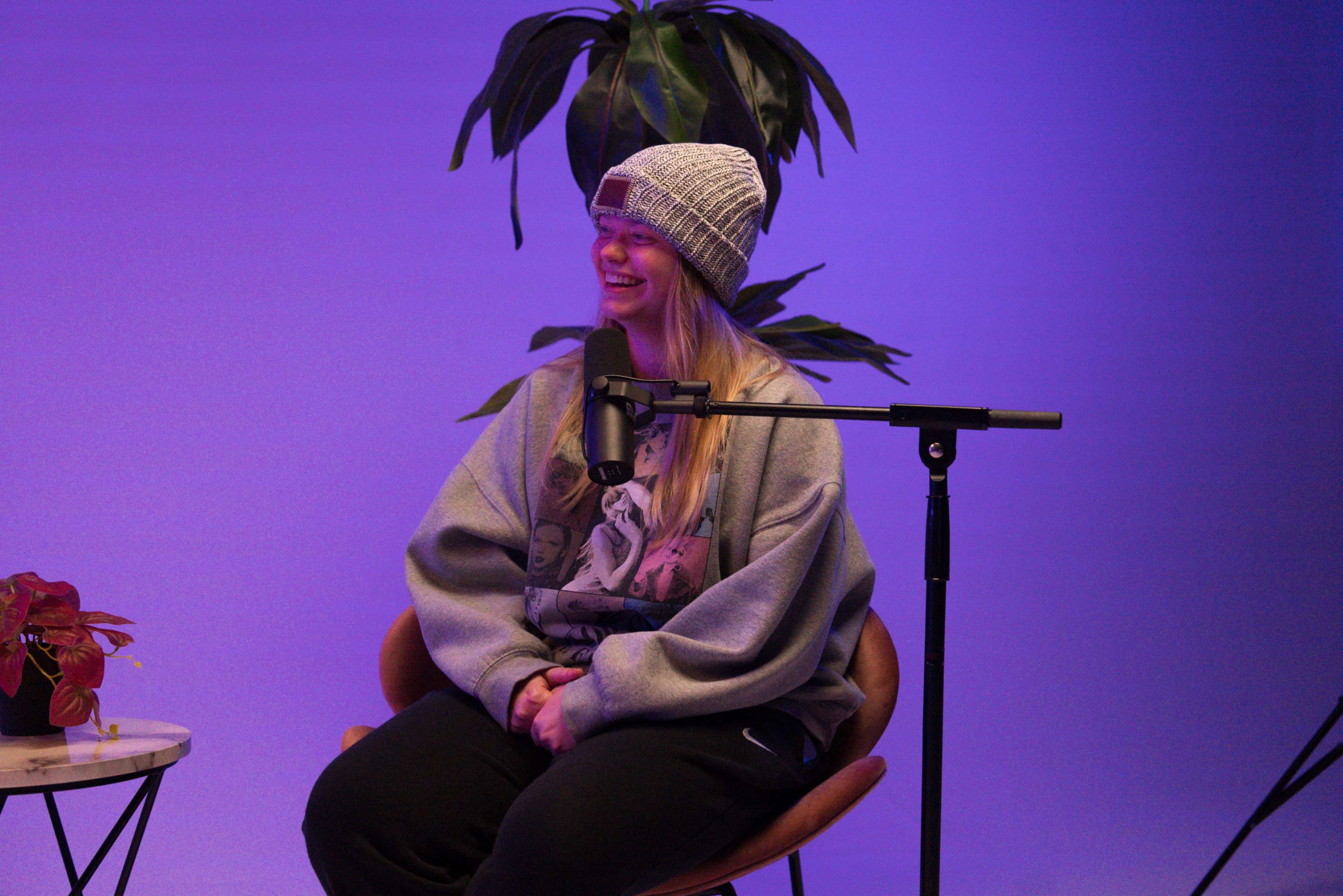 A person wearing a beanie and oversized sweatshirt sits in a chair next to a microphone and a small table with a plant in front of a purple backdrop.