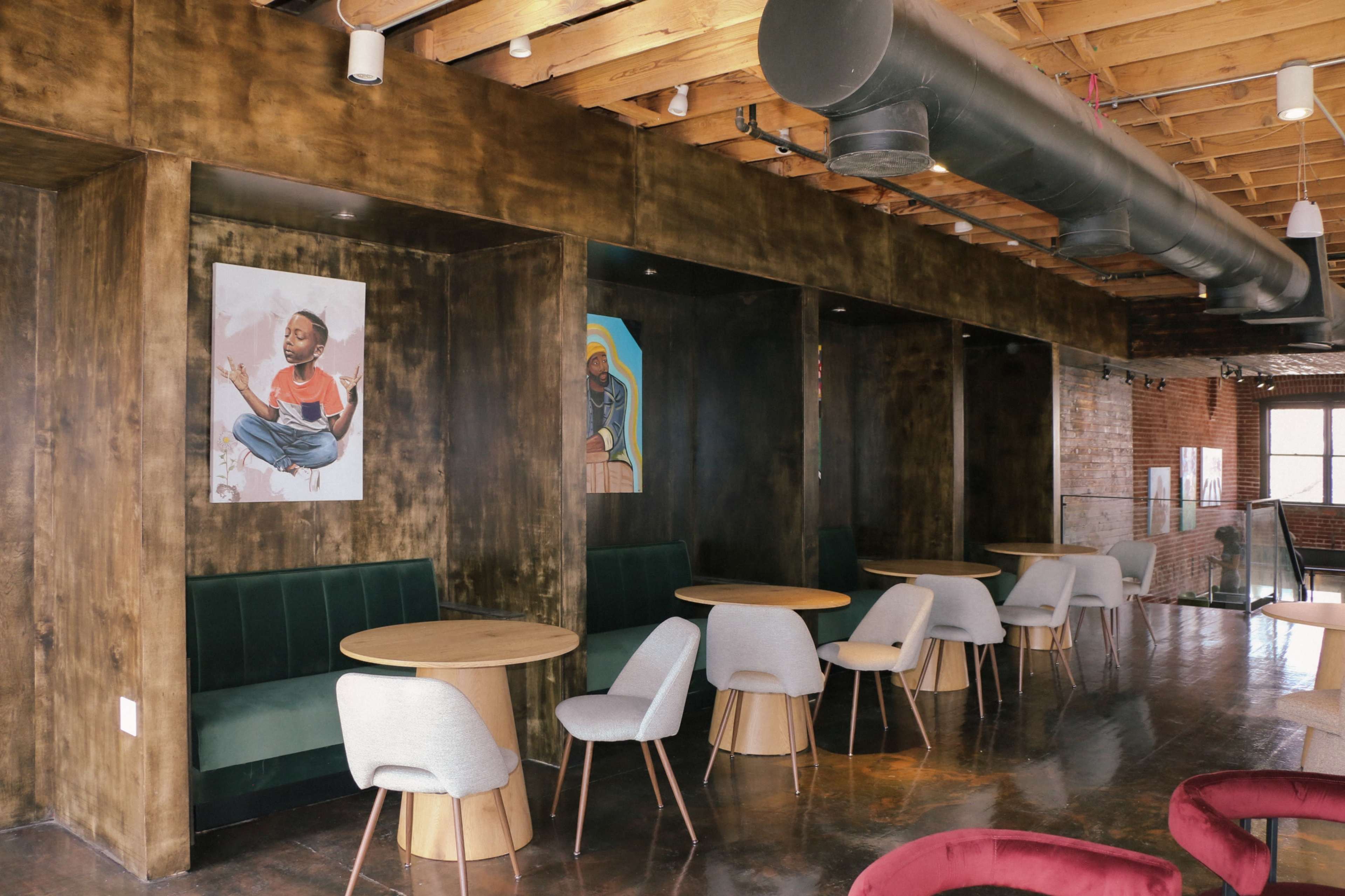 The image shows a modern café interior with wooden walls, circular tables, and upholstered chairs, featuring artwork on the walls.