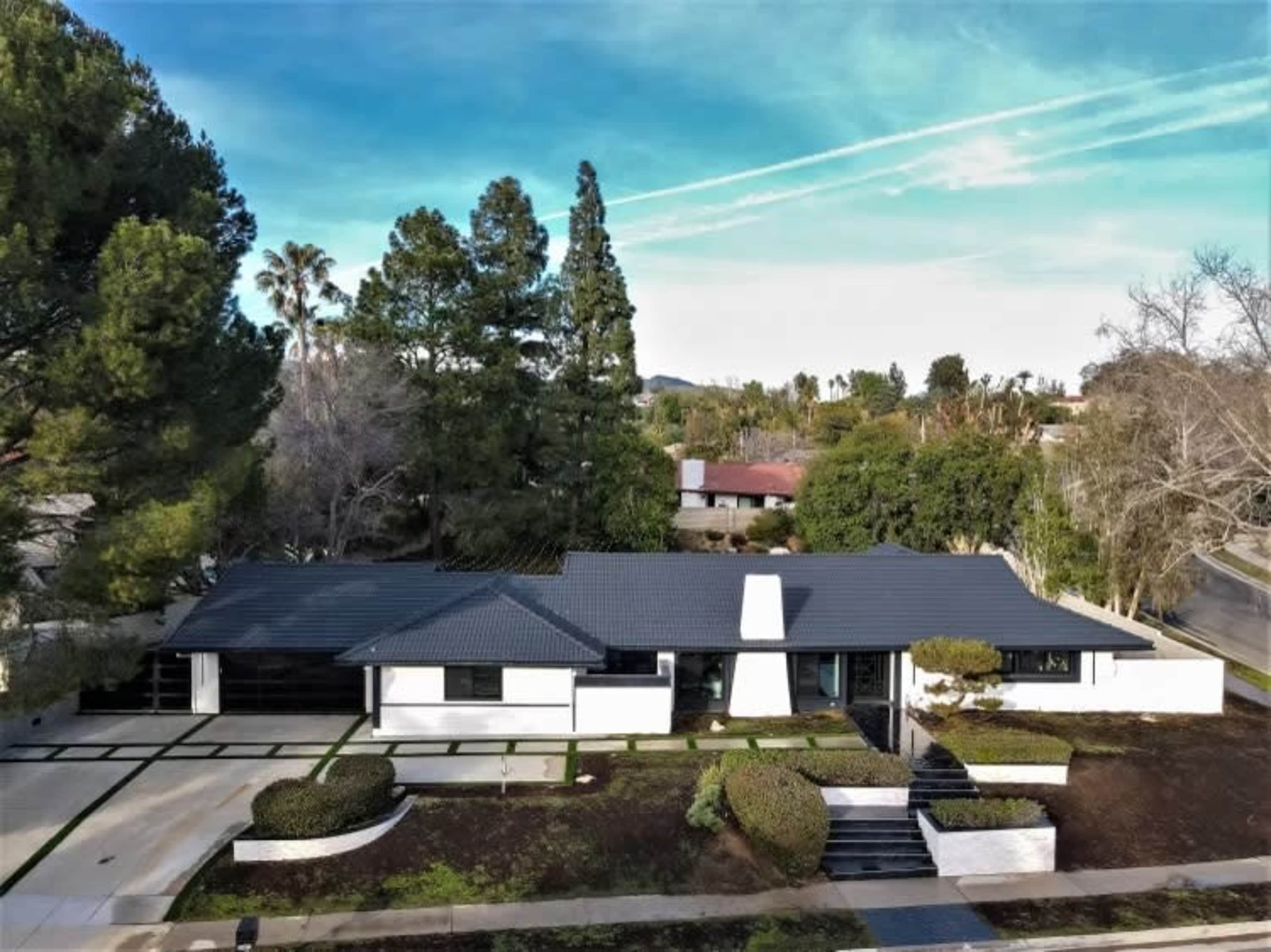 A single-story modern house with a dark roof and white exterior is surrounded by neatly landscaped greenery and trees.