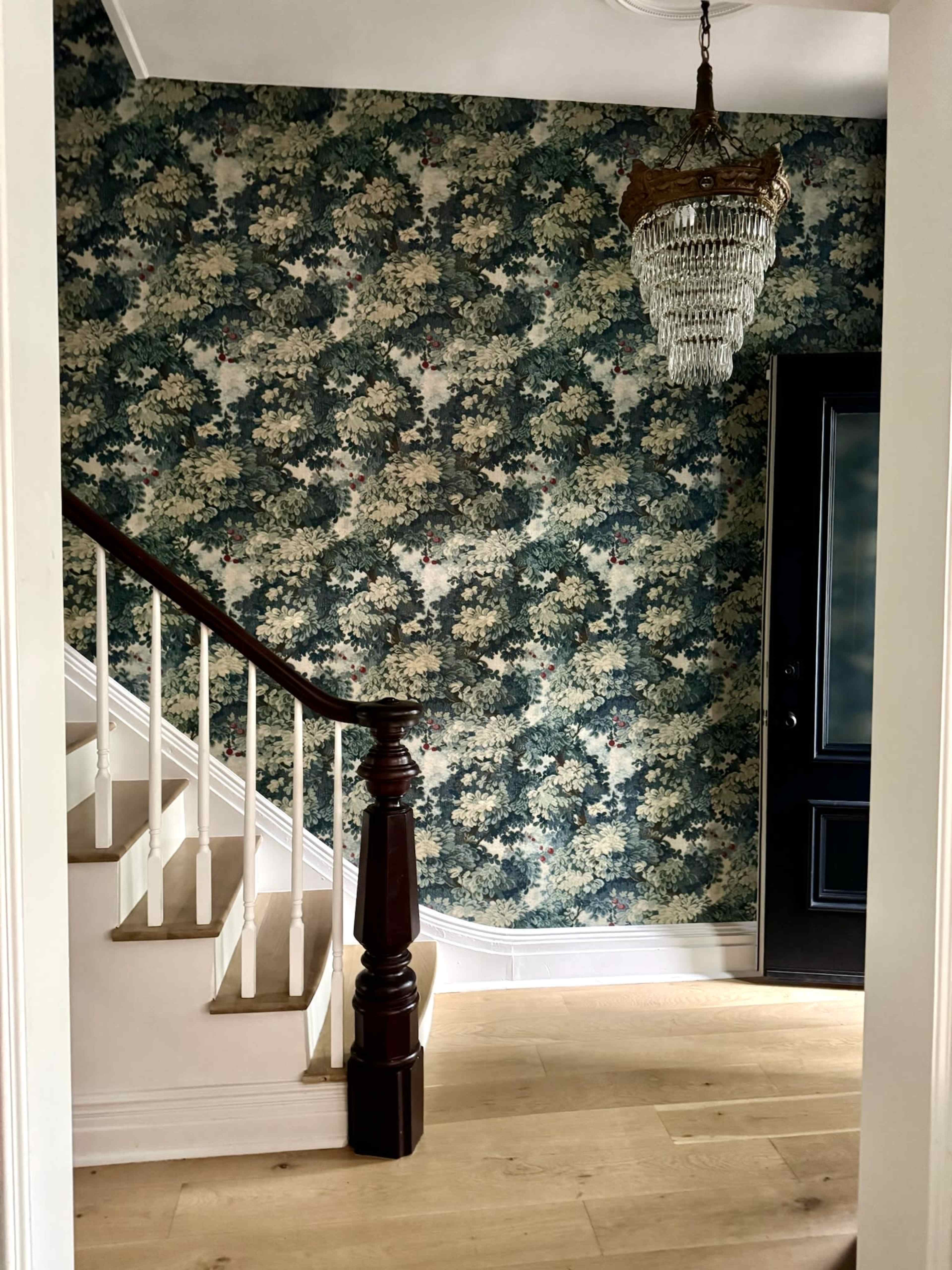 The image shows a staircase with a wooden banister, leading up against a wallpapered wall featuring a floral pattern, with a crystal chandelier hanging above and a dark door visible on the right.