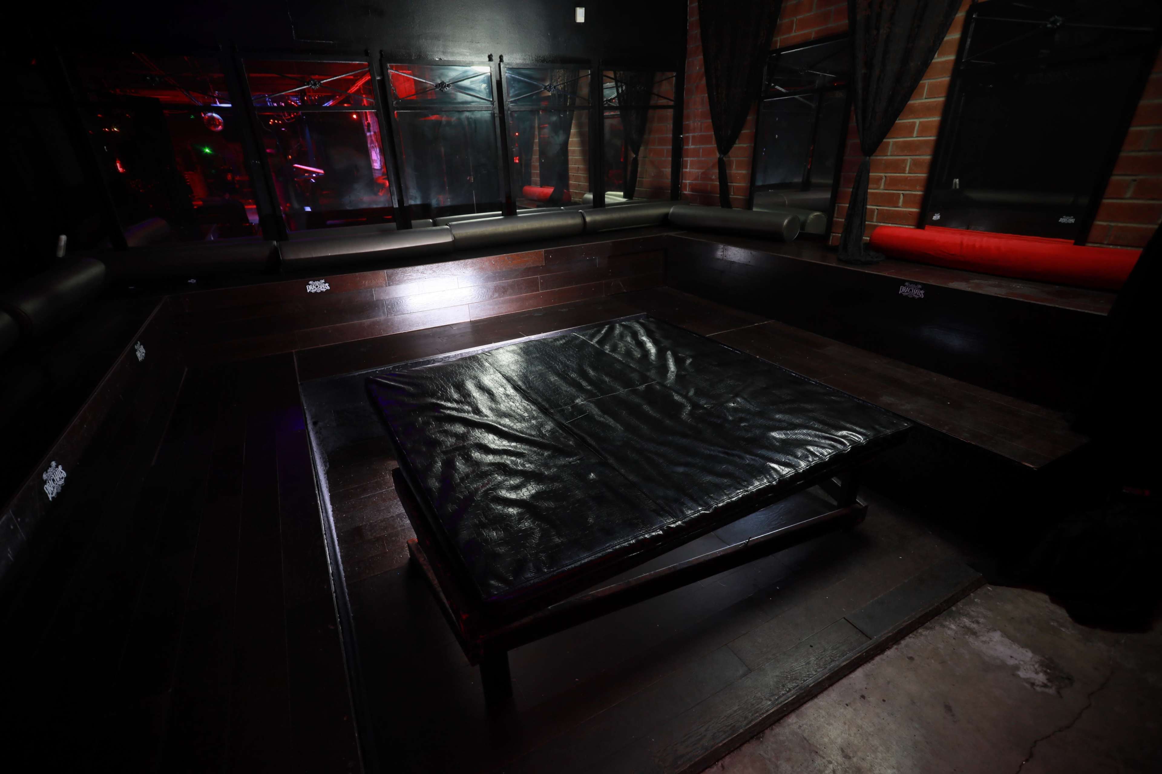 The image shows an empty seating area with a large black padded platform in a dimly lit room featuring dark walls and red ambient lighting.