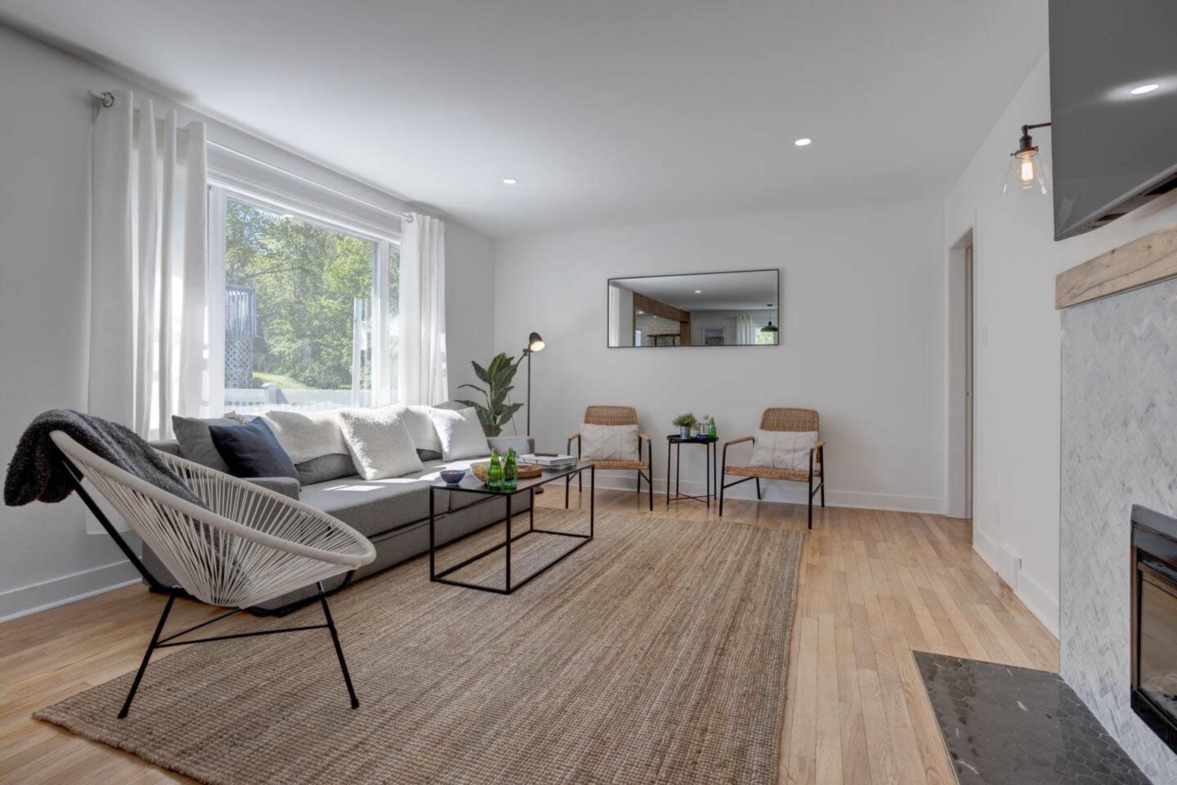 The room features a modern living space with a sofa, two chairs, a coffee table, and a fireplace, illuminated by natural light from a large window.
