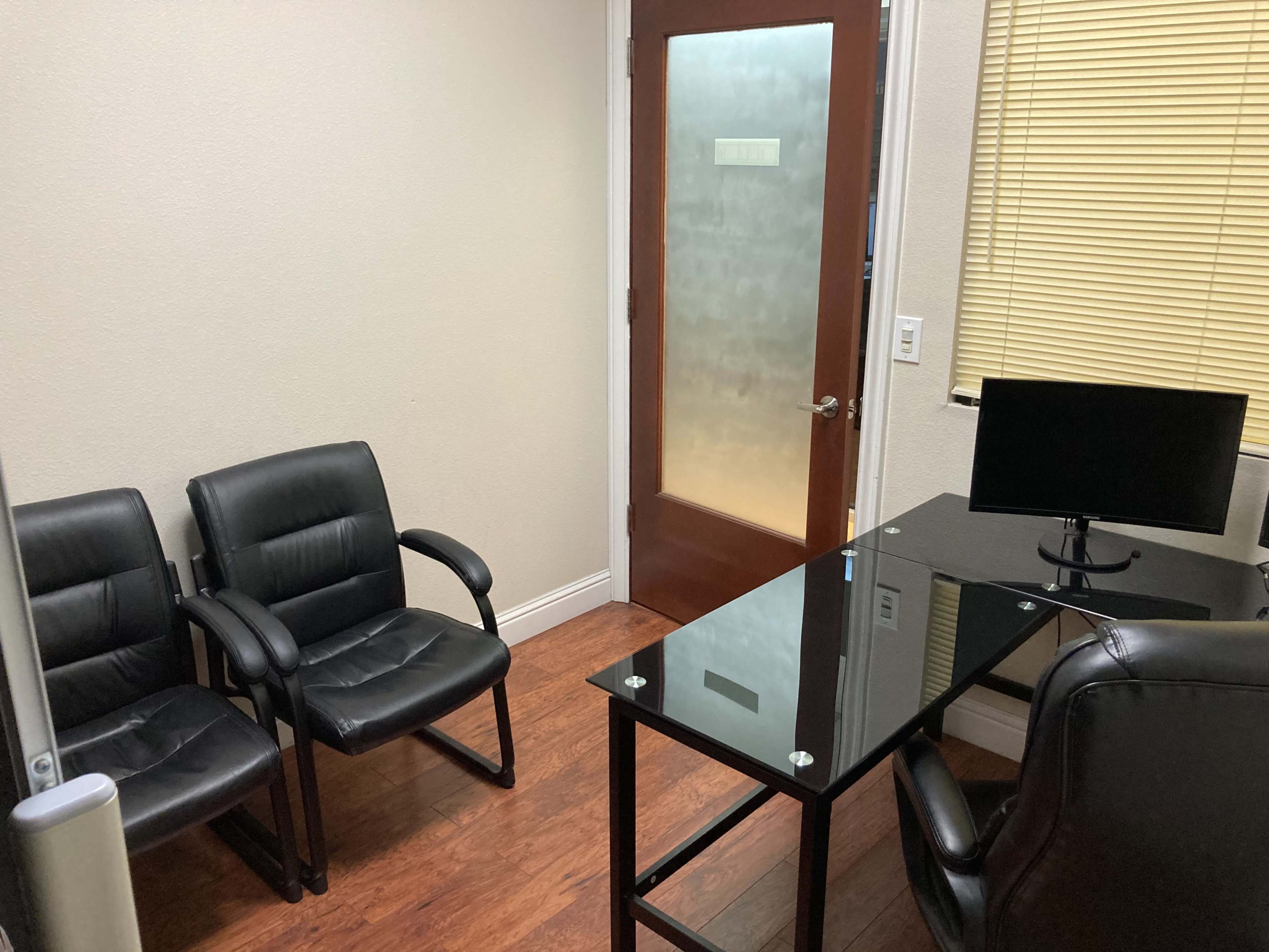 A small office features two black chairs, a glass-topped desk with a computer monitor, and a wooden door with frosted glass.