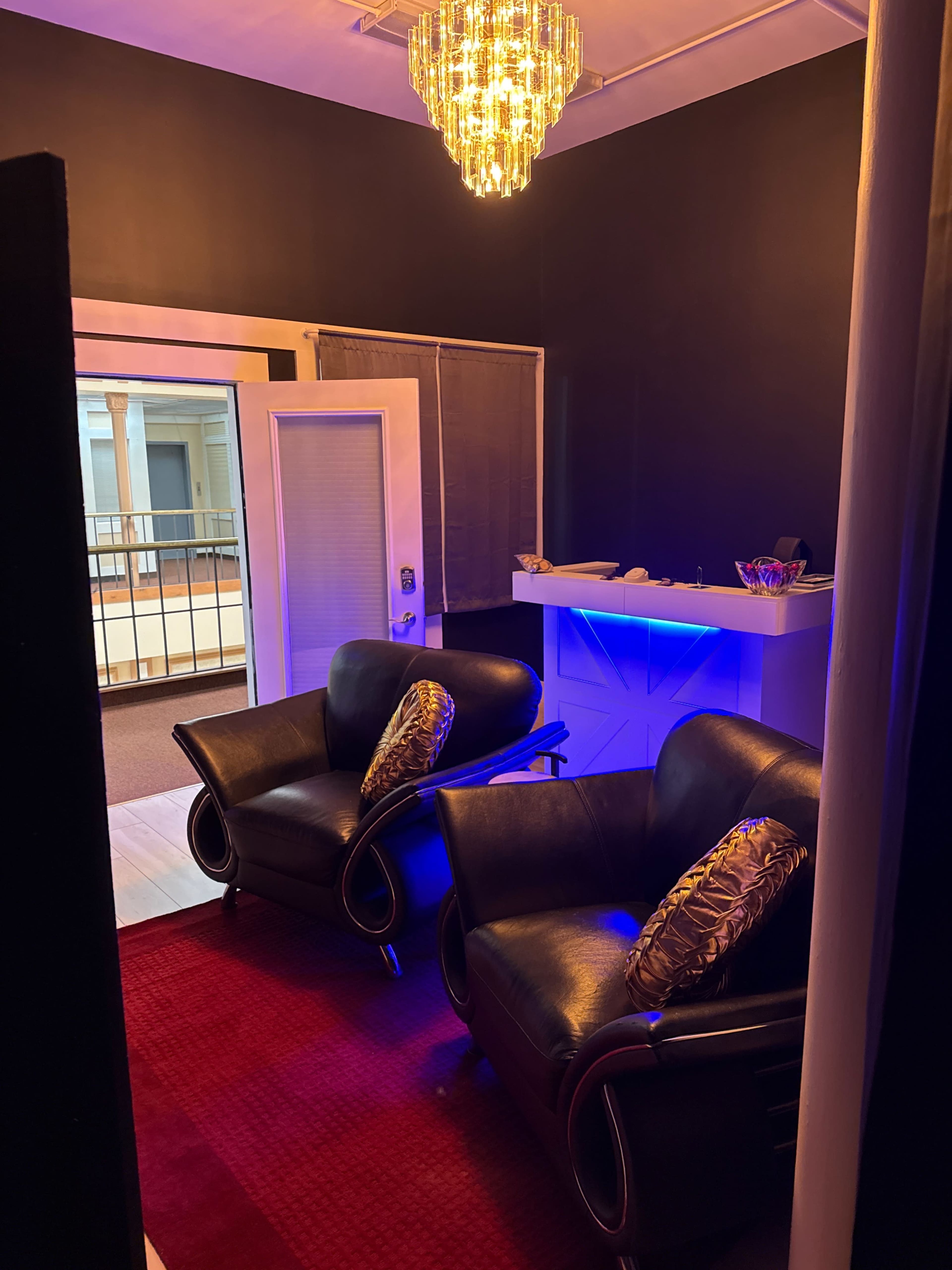 A modern lounge area features two black chairs with decorative pillows and a red rug, illuminated by a chandelier and blue lighting.