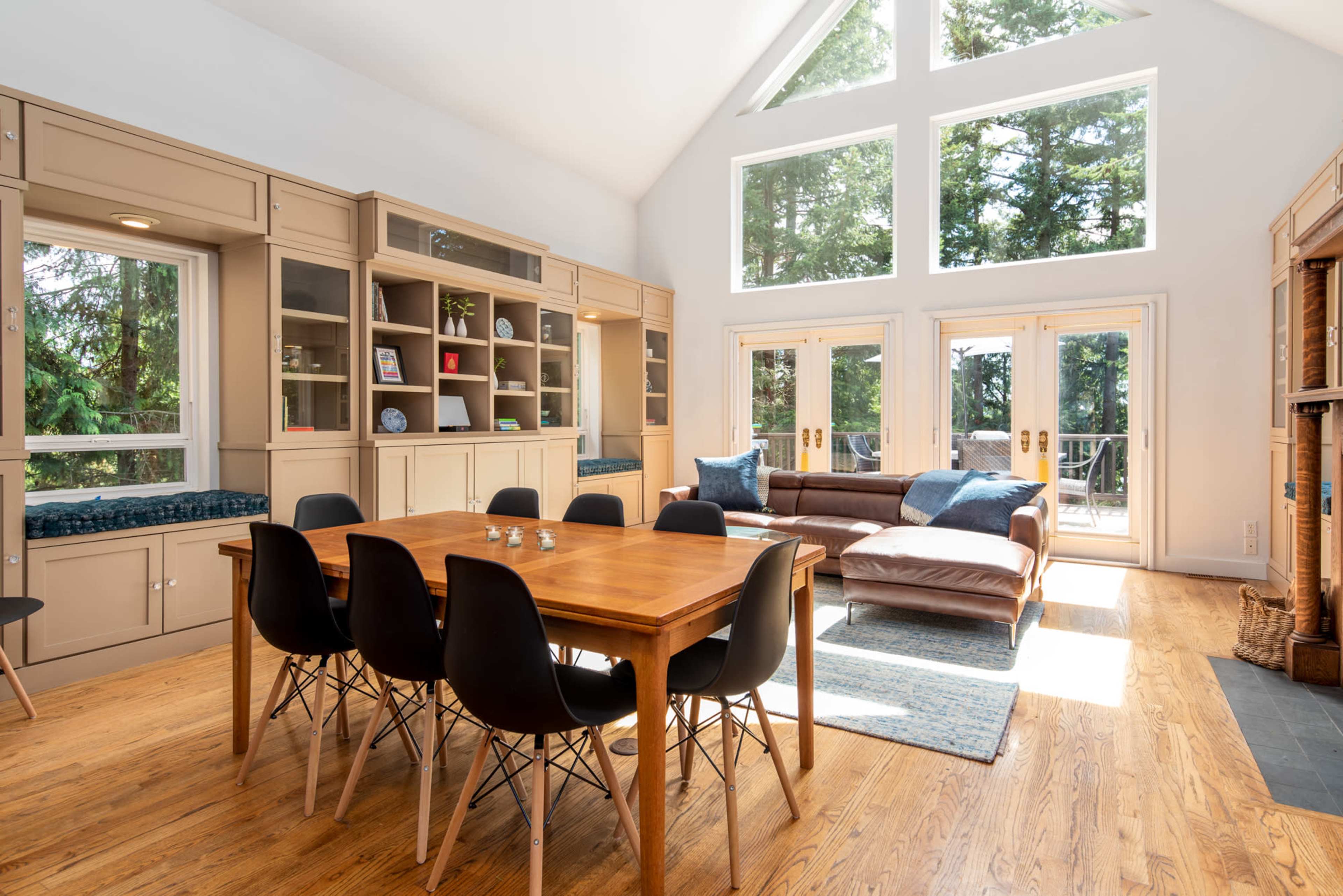 A bright living room features a wooden dining table surrounded by six black chairs, with large windows allowing natural light and views of the trees outside.