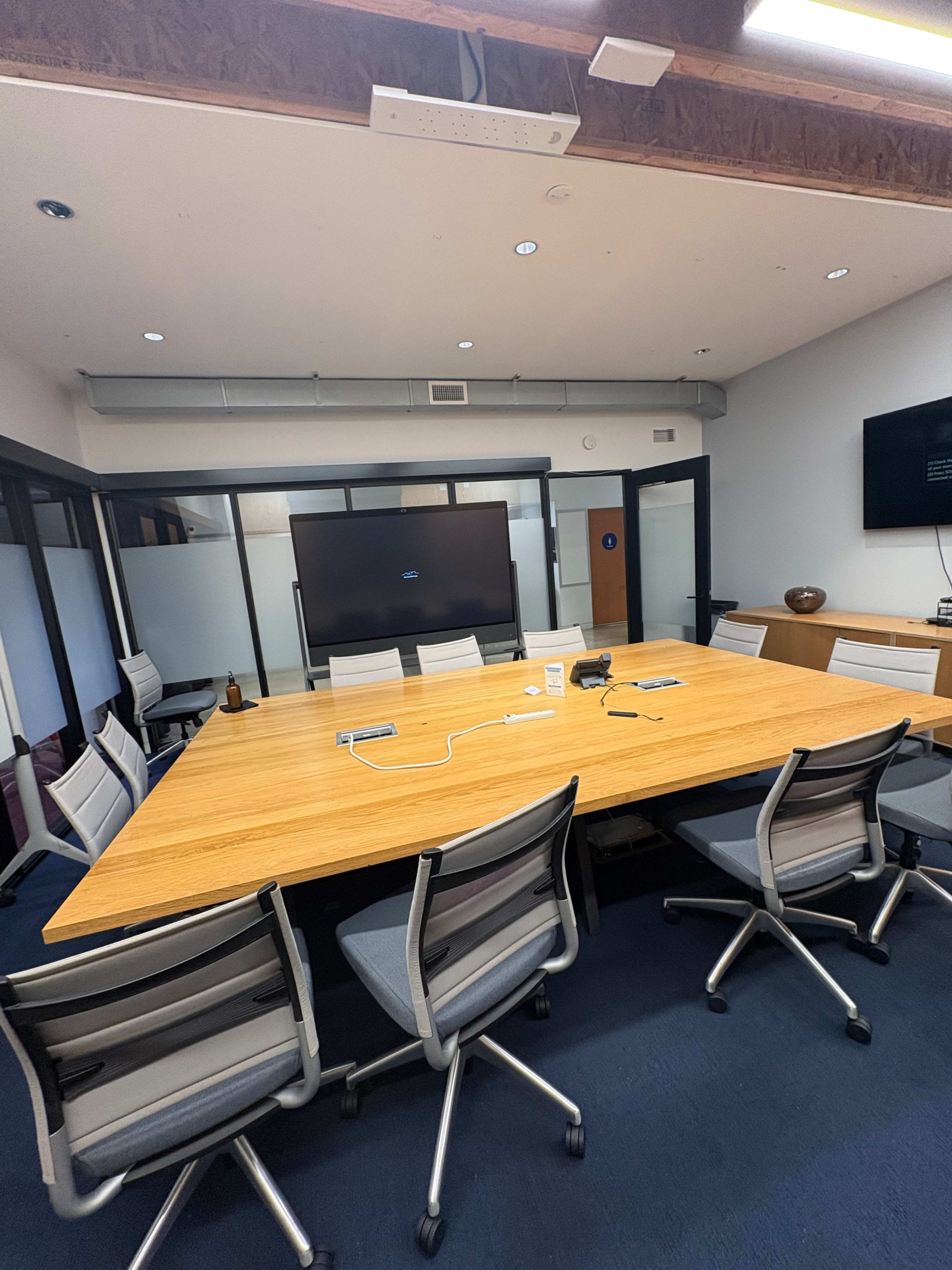 The image shows a modern conference room with a large wooden table surrounded by gray office chairs and a display screen on the wall.