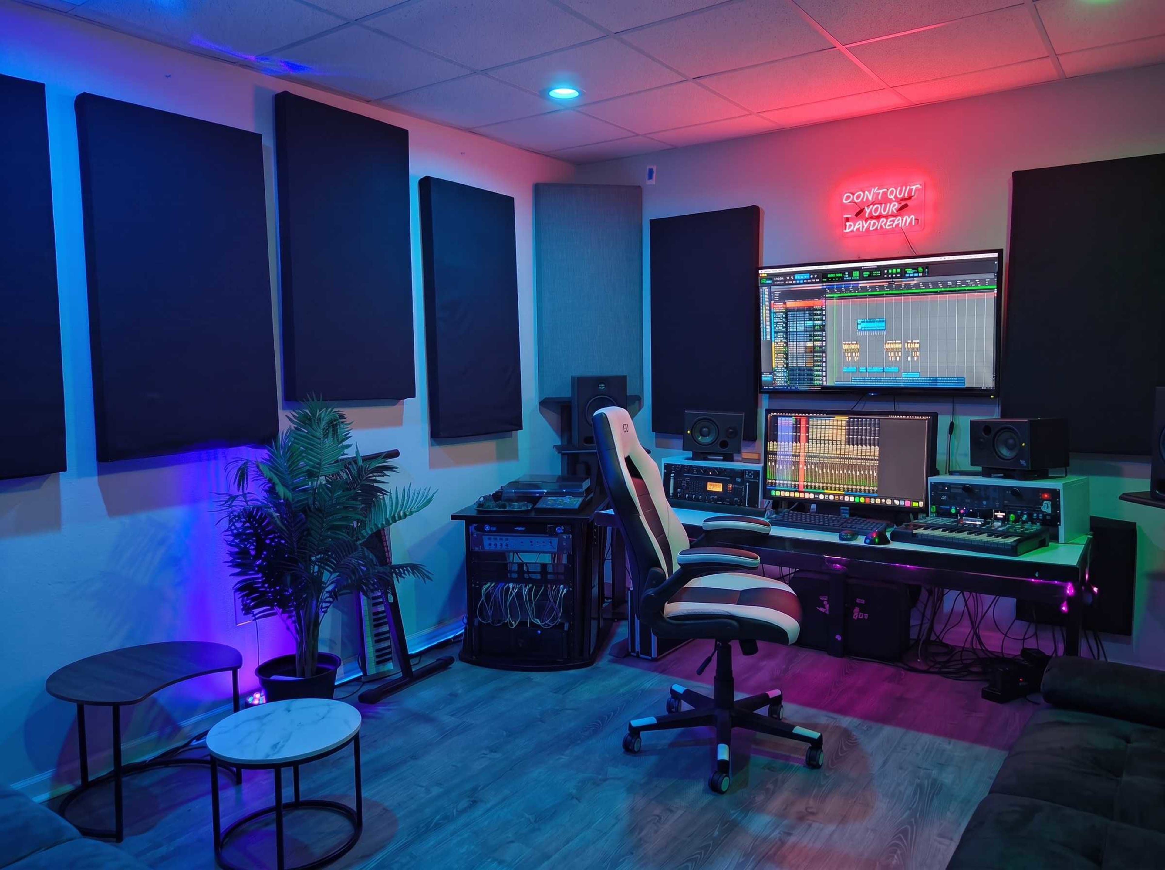 A music studio is set up with sound-absorbing panels, a computer workstation, and various audio equipment, illuminated by colorful lights.