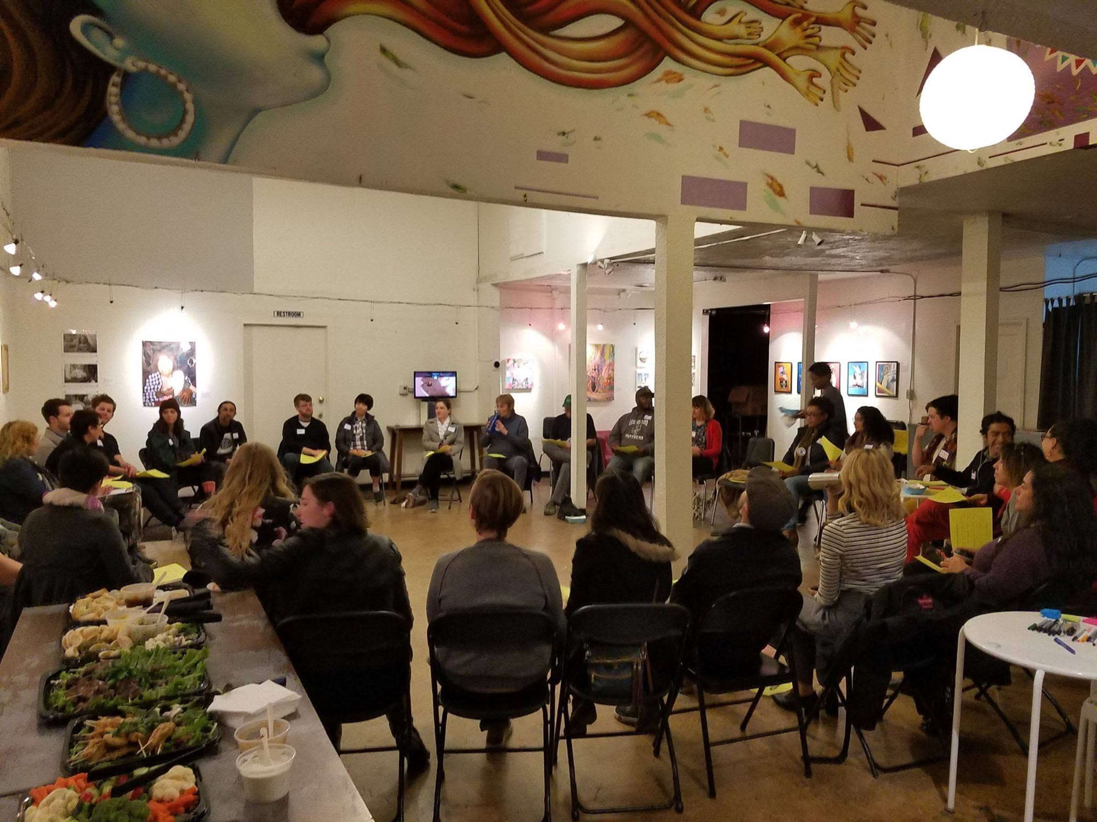 A diverse group of people sits in a circle in a spacious room adorned with colorful wall art, engaging in discussion while a table with food is set nearby.