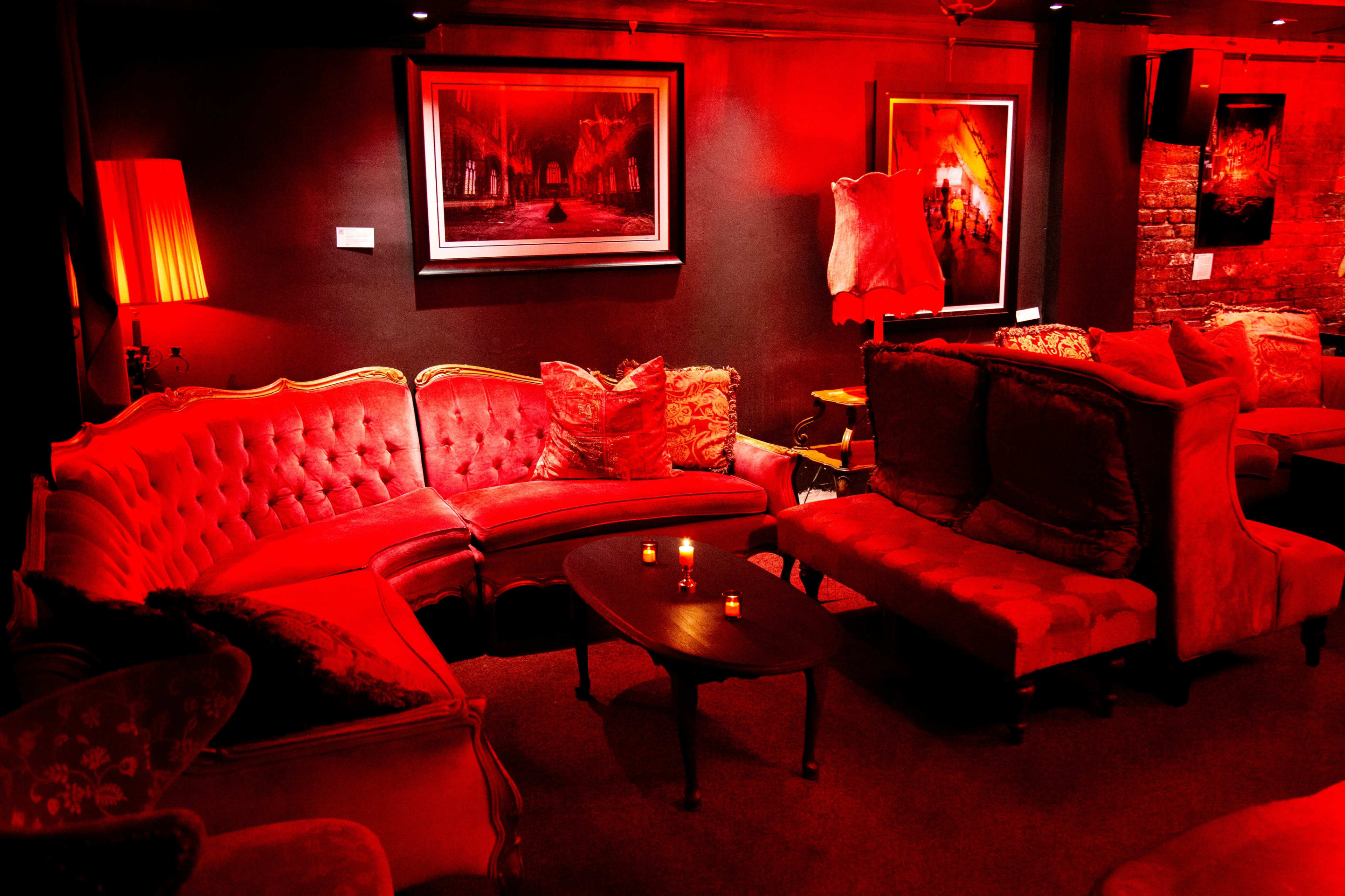 A lounge area with red lighting, featuring a plush red sectional sofa, a small coffee table, and framed artworks on the walls.