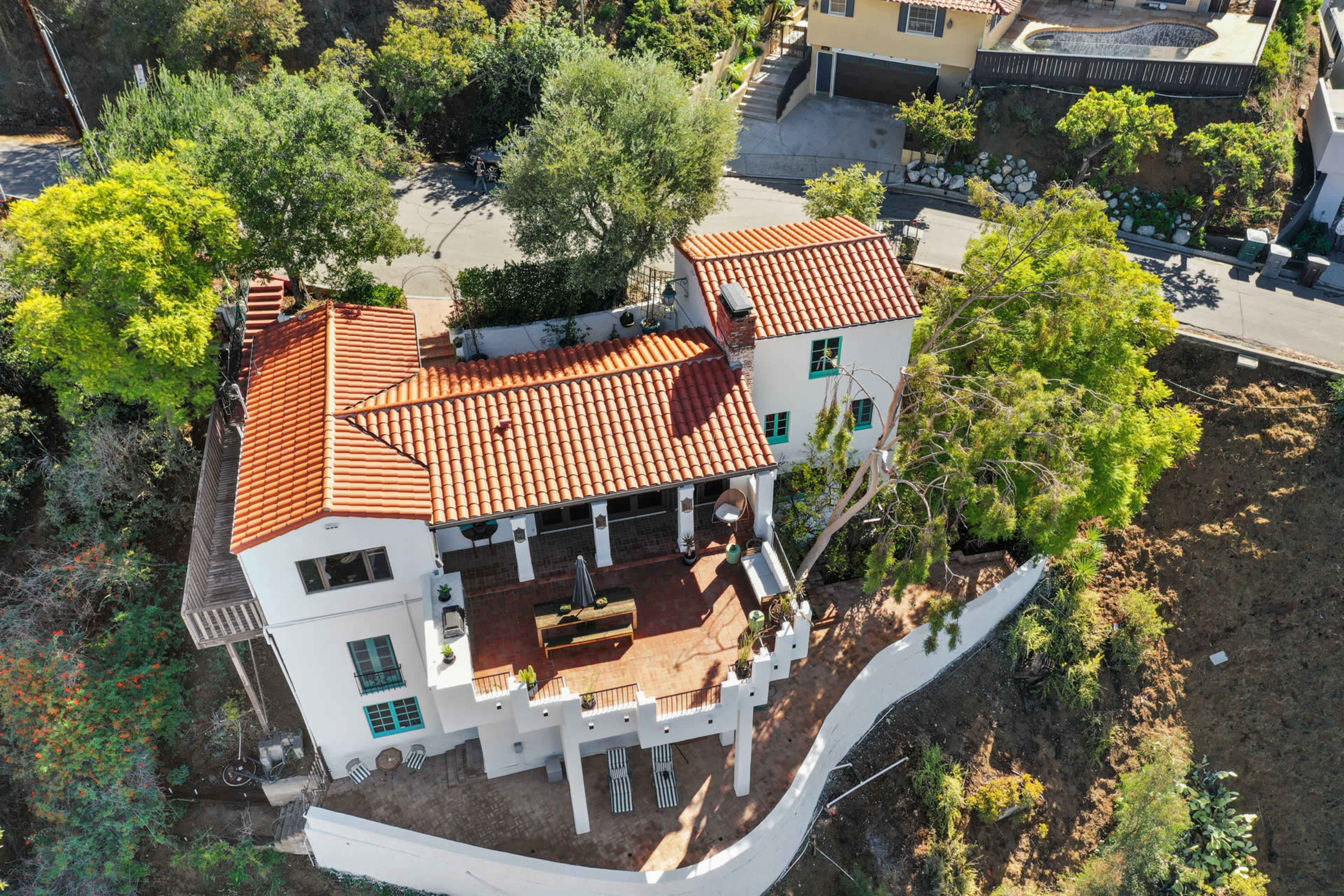 A two-story white house with a red tile roof, situated on a hillside surrounded by trees, with a patio area and a road visible in the background.