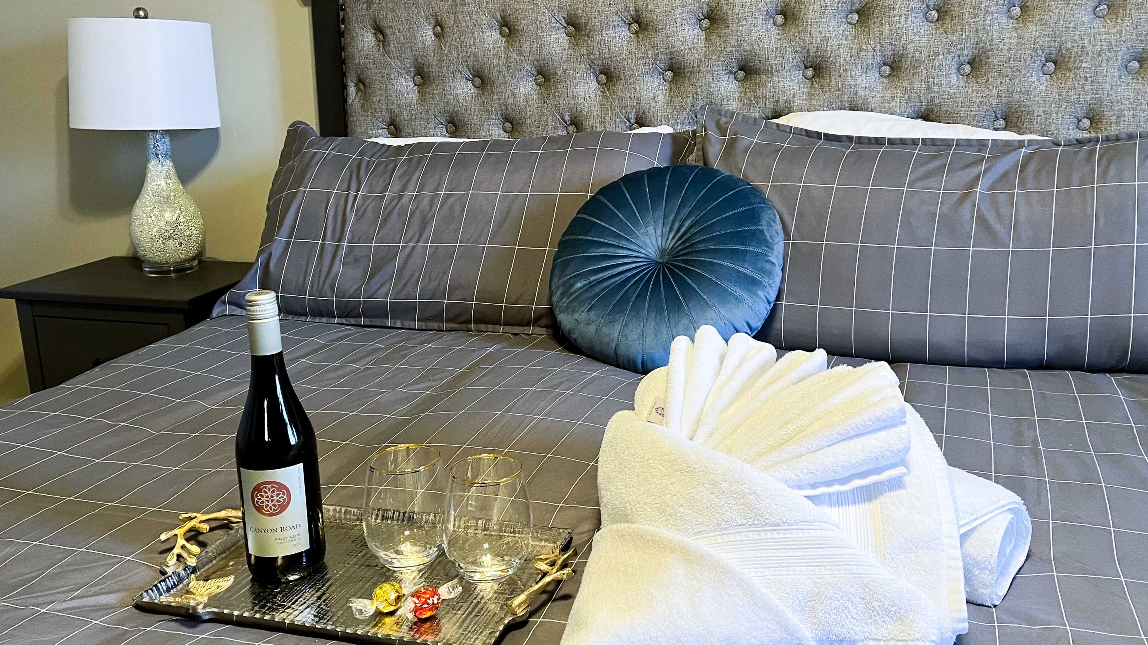 A neatly arranged bed features gray plaid linens, a decorative blue pillow, a bottle of wine, two glasses, and folded towels on a tray.