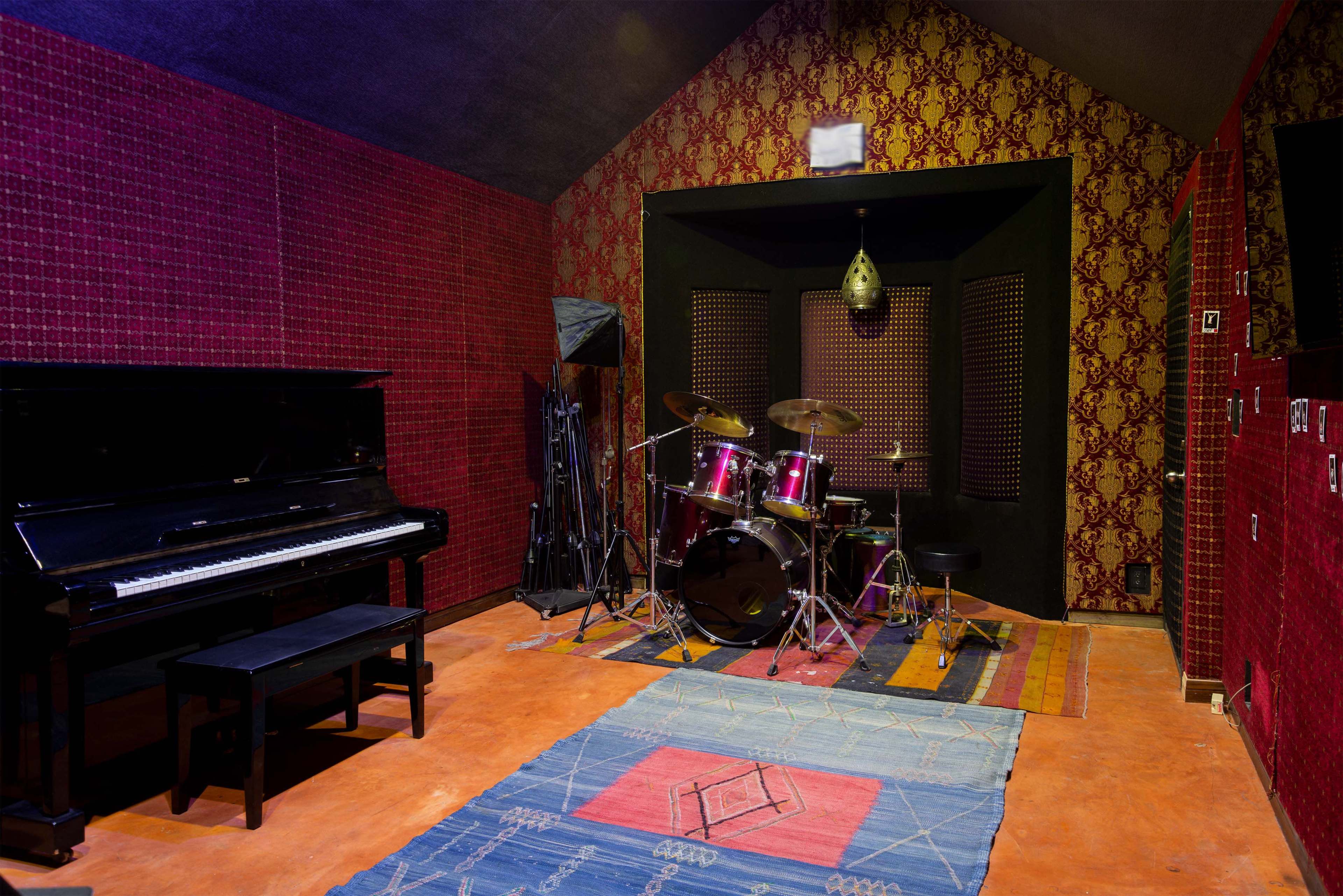 A music studio featuring a grand piano on one side and a drum set on the other, surrounded by red patterned walls.