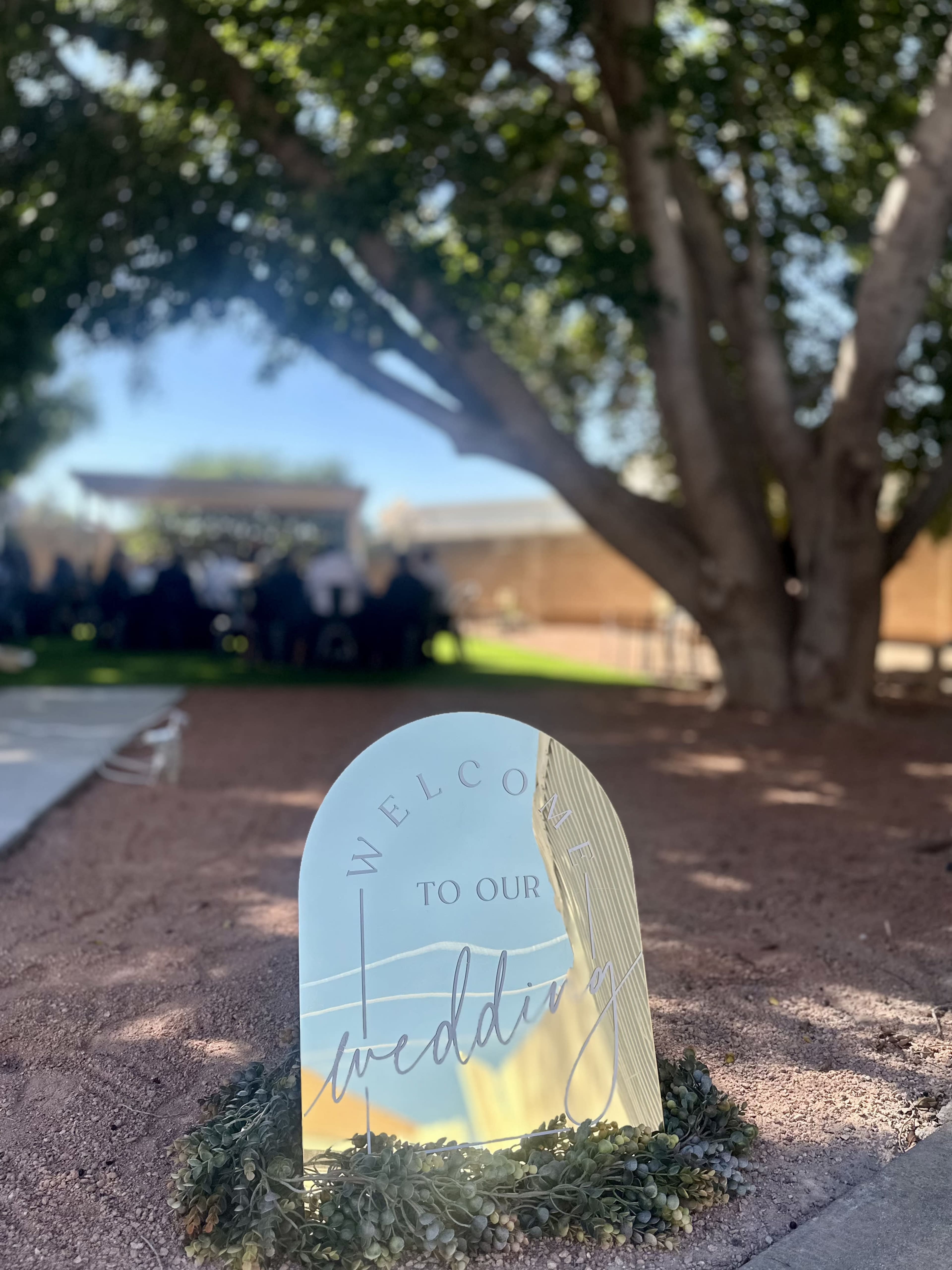 A decorative sign reading "Welcome to our wedding" stands in a garden setting, with guests seated in the background.