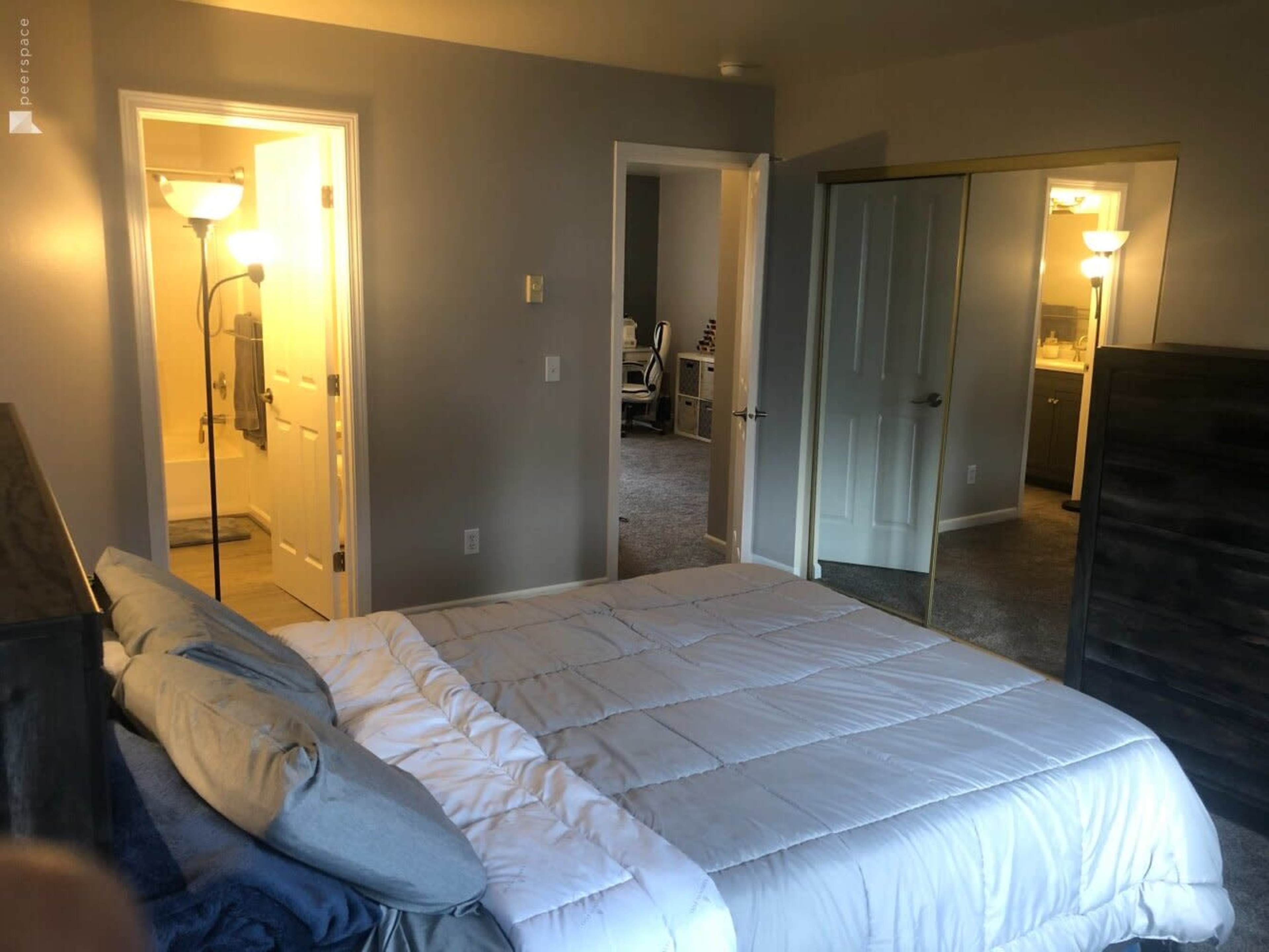 A neatly arranged bedroom features a bed with white bedding, and doorways lead to other rooms with lighted interiors.