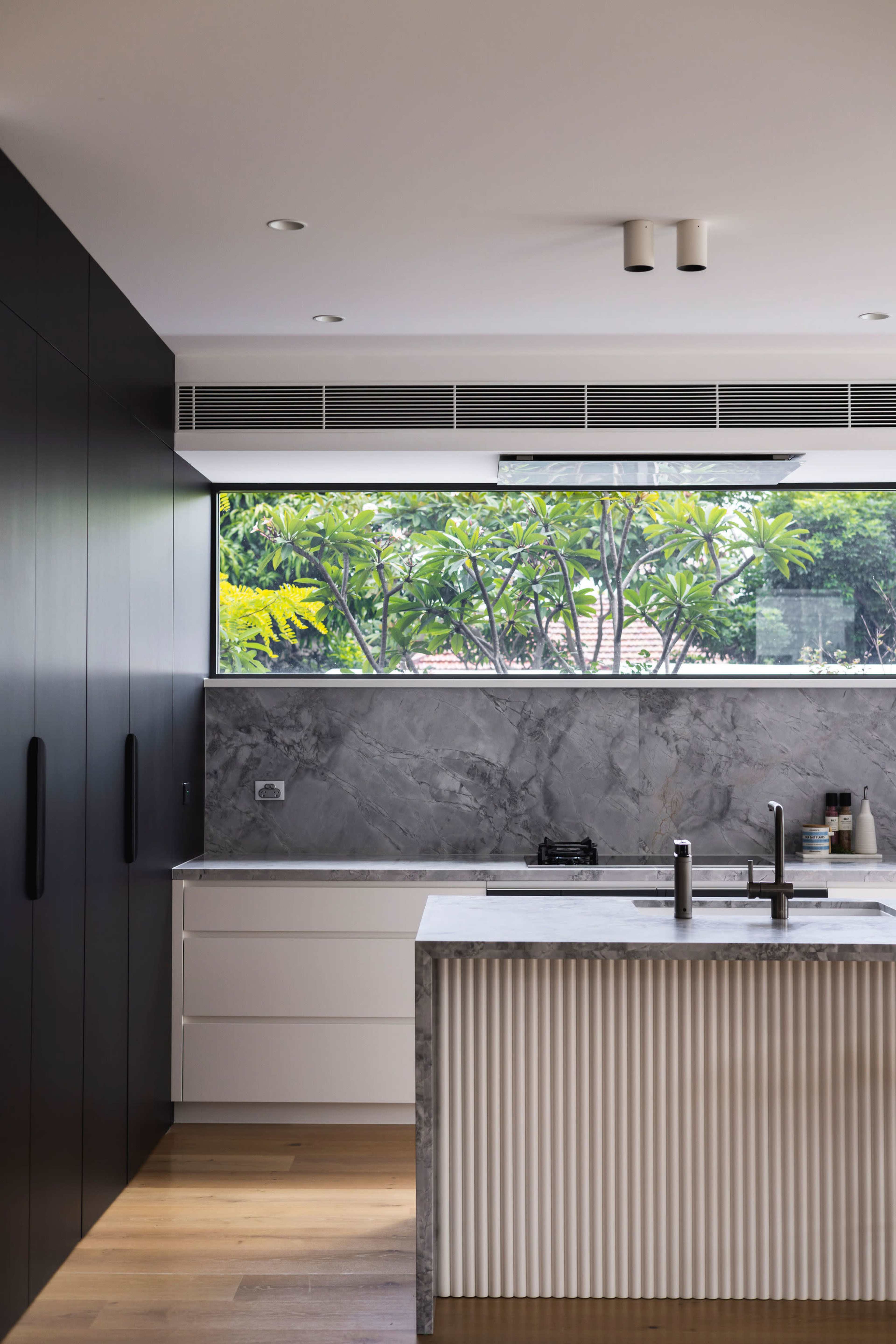 A modern kitchen with a central island, featuring a marble countertop and a large window overlooking greenery.