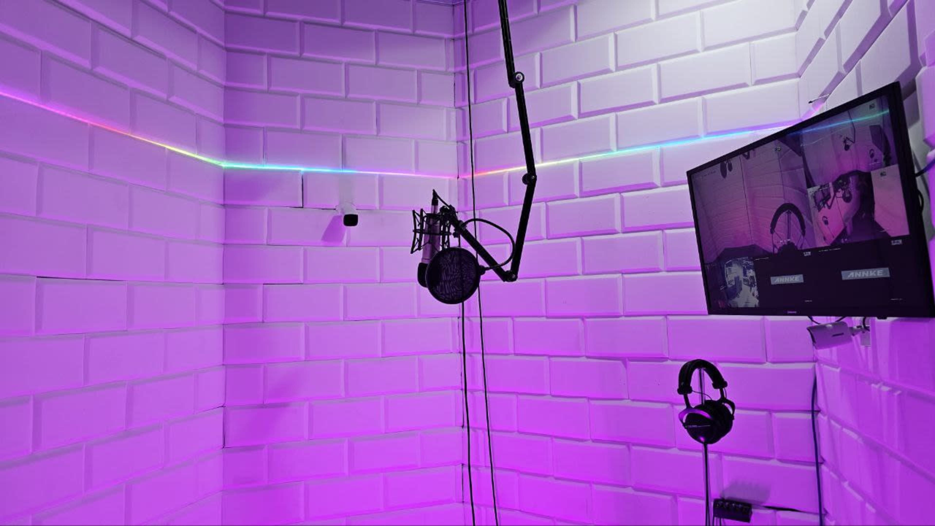 A soundproof recording booth with a microphone on a stand, a monitor displaying audio production software, and a pair of headphones hanging nearby, all illuminated by purple lighting.