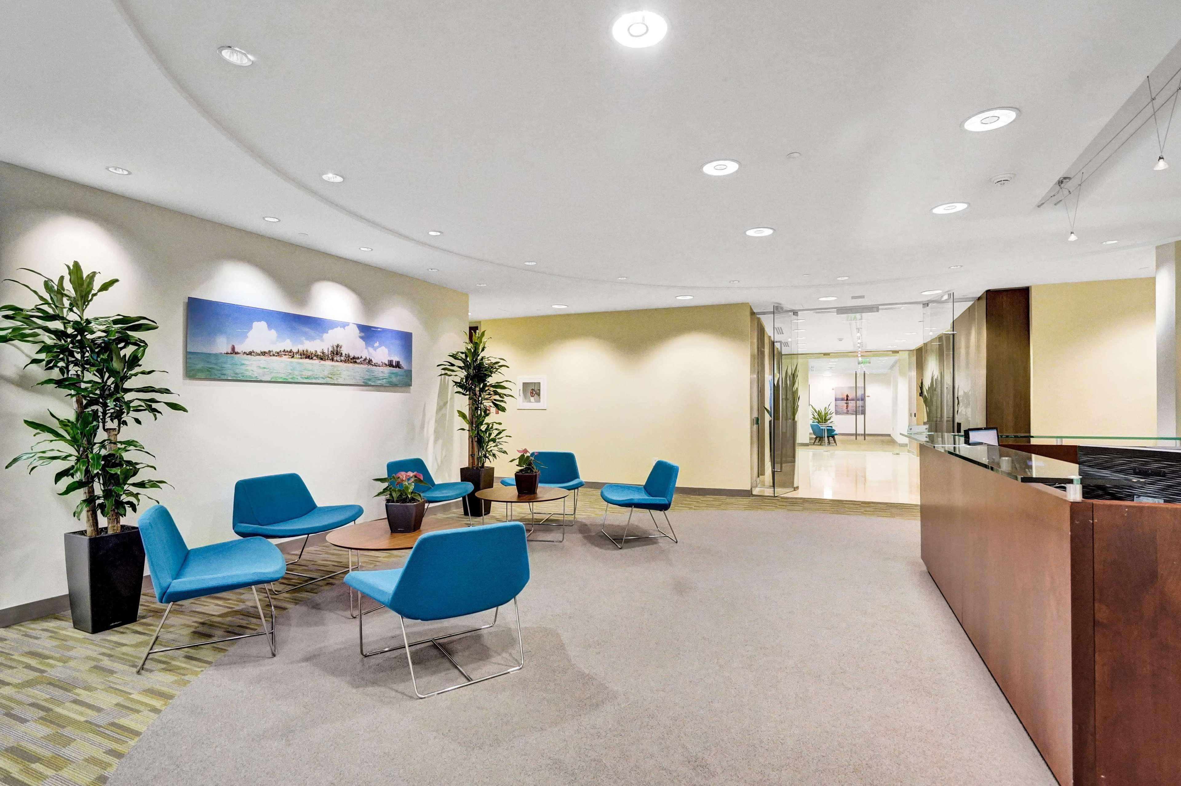 A modern office reception area featuring blue chairs, small potted plants, and a front desk with a large wall art display.