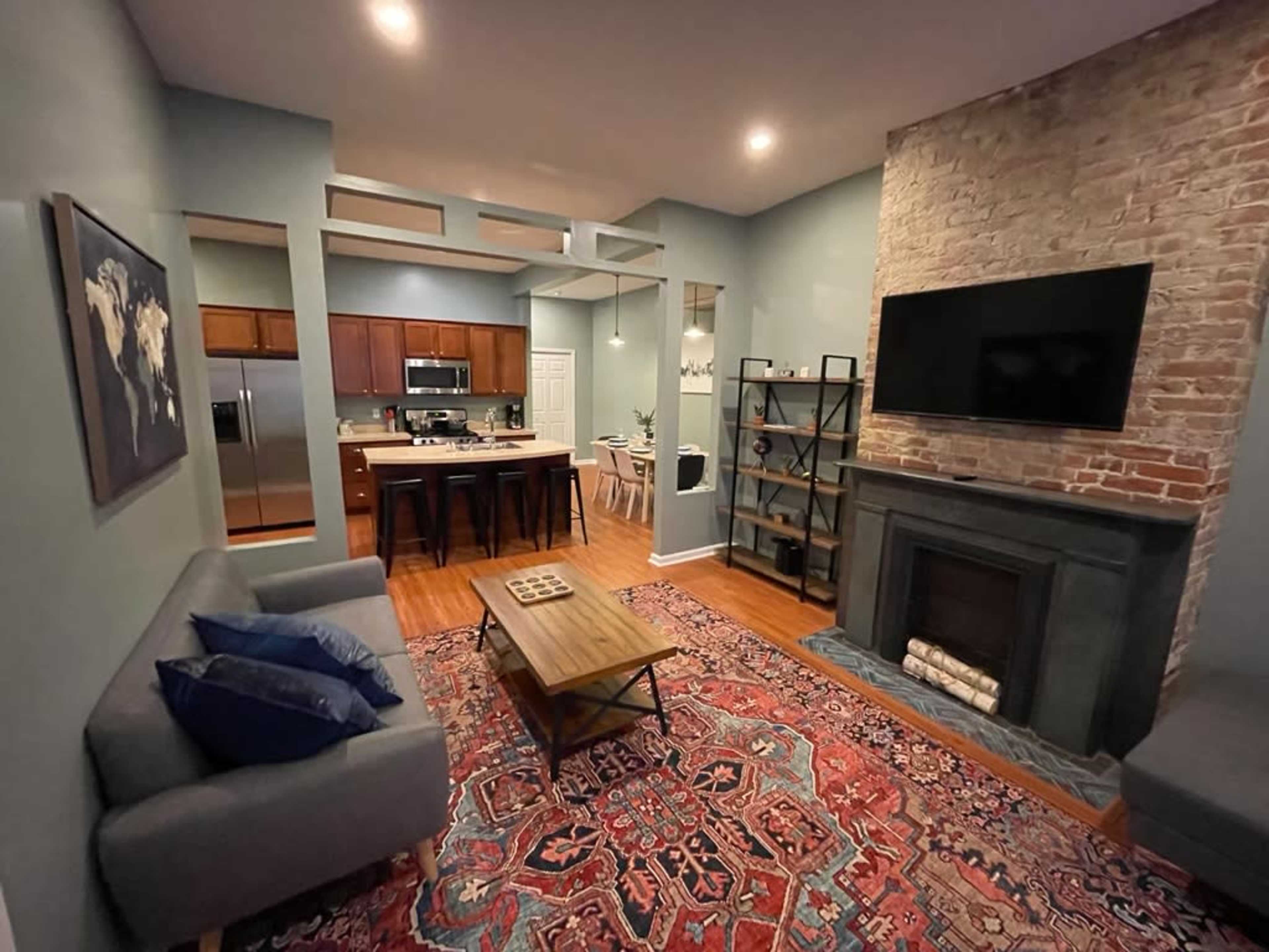 A contemporary living room with a brick fireplace, a gray sofa, a wooden coffee table, and a wall-mounted TV, accompanied by a kitchen area in the background.