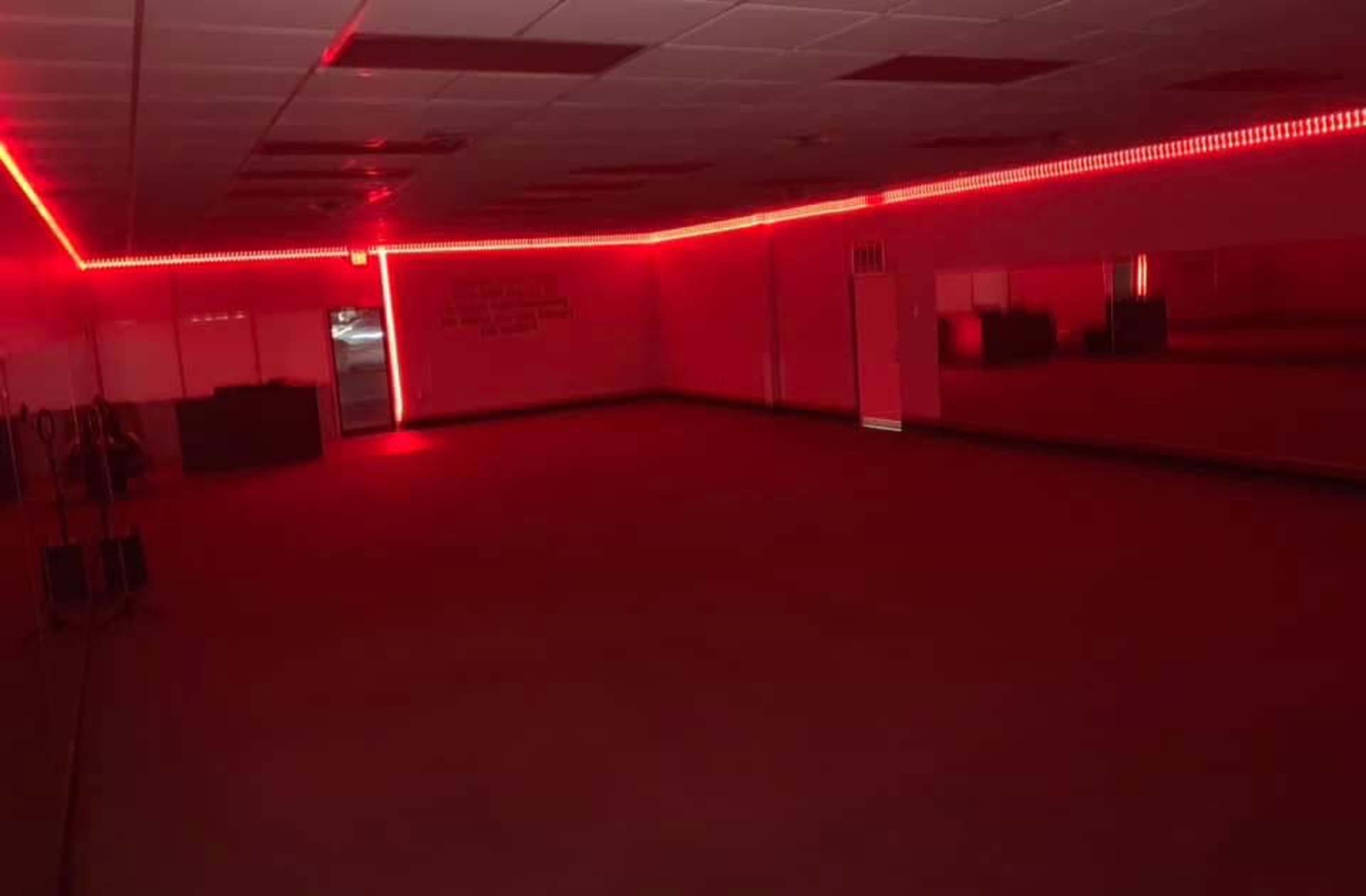 An empty room illuminated by red LED lights, featuring a large mirror on one wall and a door visible at the far end.