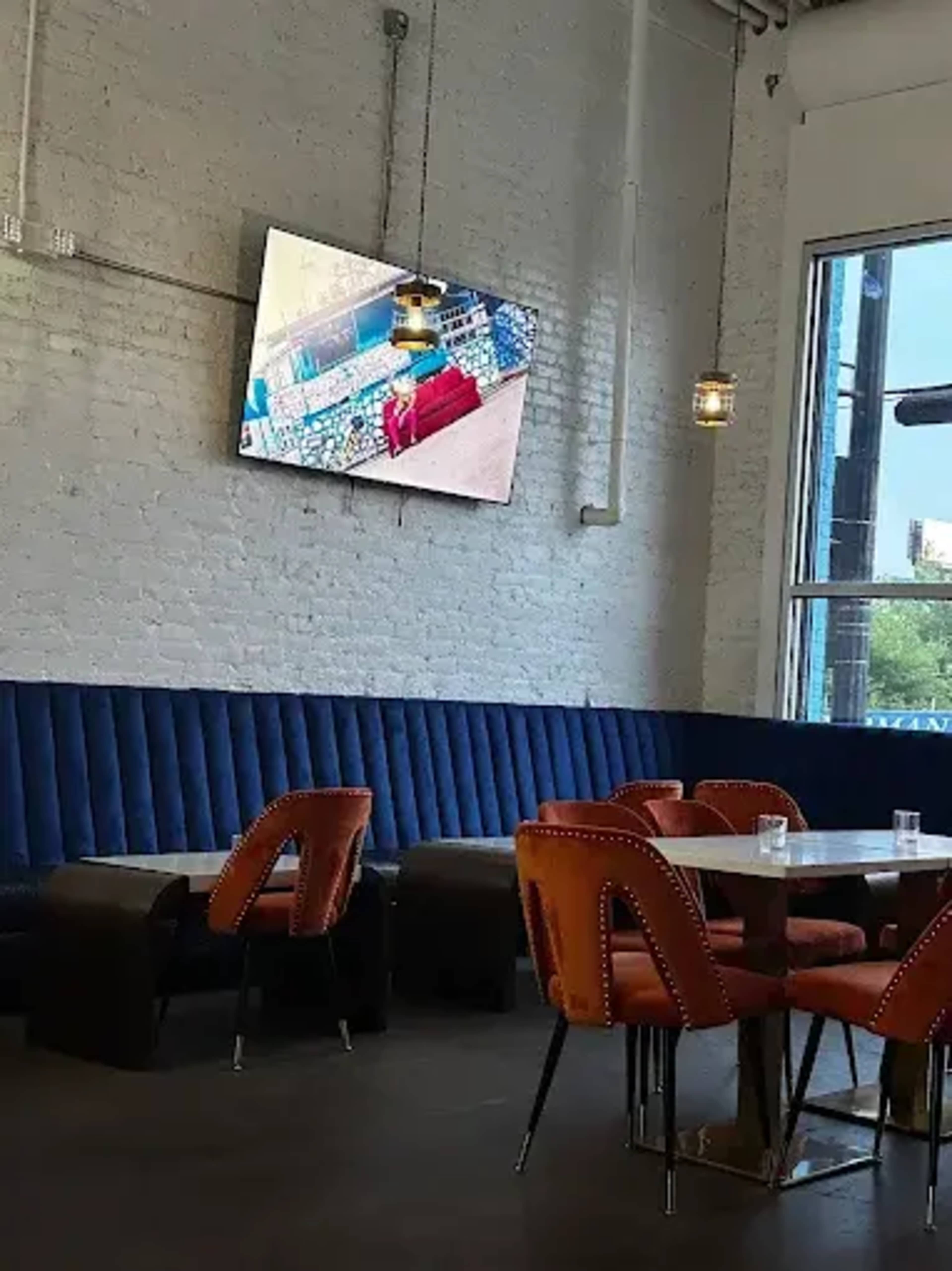 The image shows a modern restaurant interior featuring a blue upholstered banquette, orange chairs, wooden tables, and a flat-screen TV mounted on the wall.